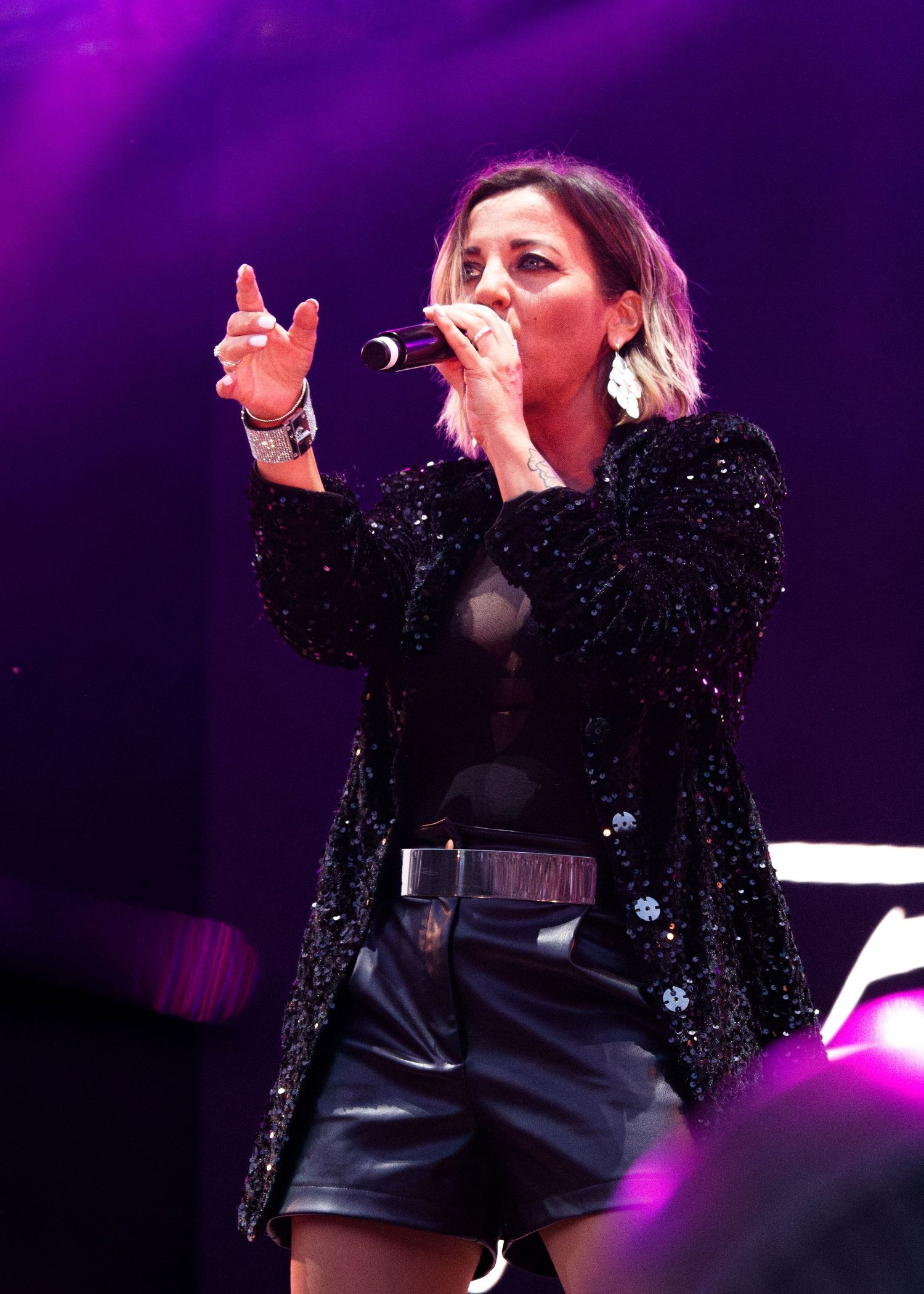 A performer in a sparkly black blazer and shorts sings on stage under dramatic purple lighting.