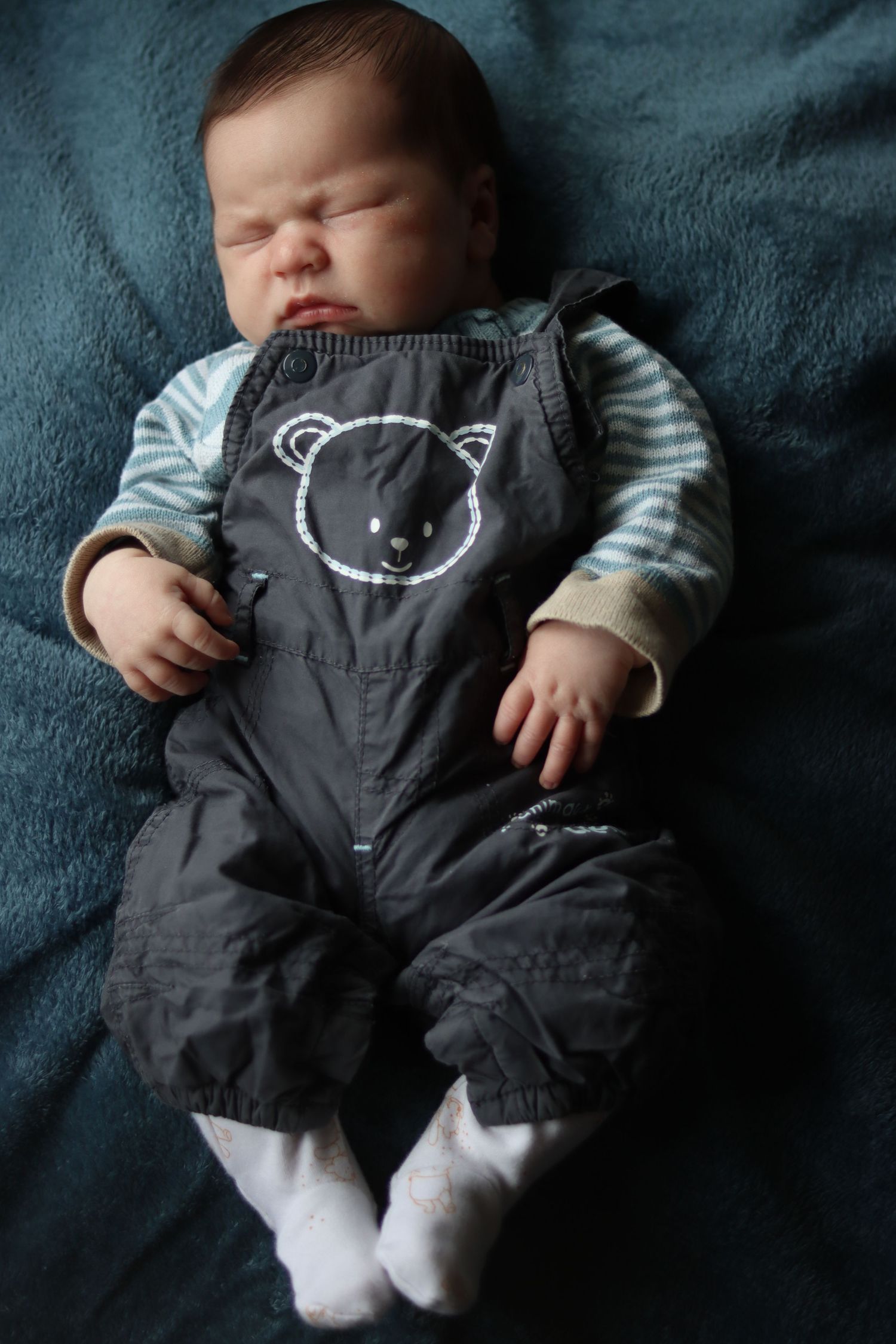 Sleeping baby wearing grey overalls with teddy bear design against dark background.