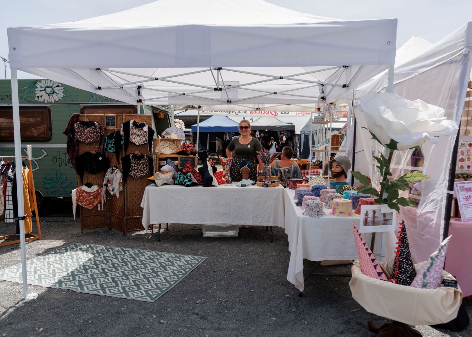 Outdoor market vendor booth with white canopy displaying clothing, accessories, and handmade goods on tables.