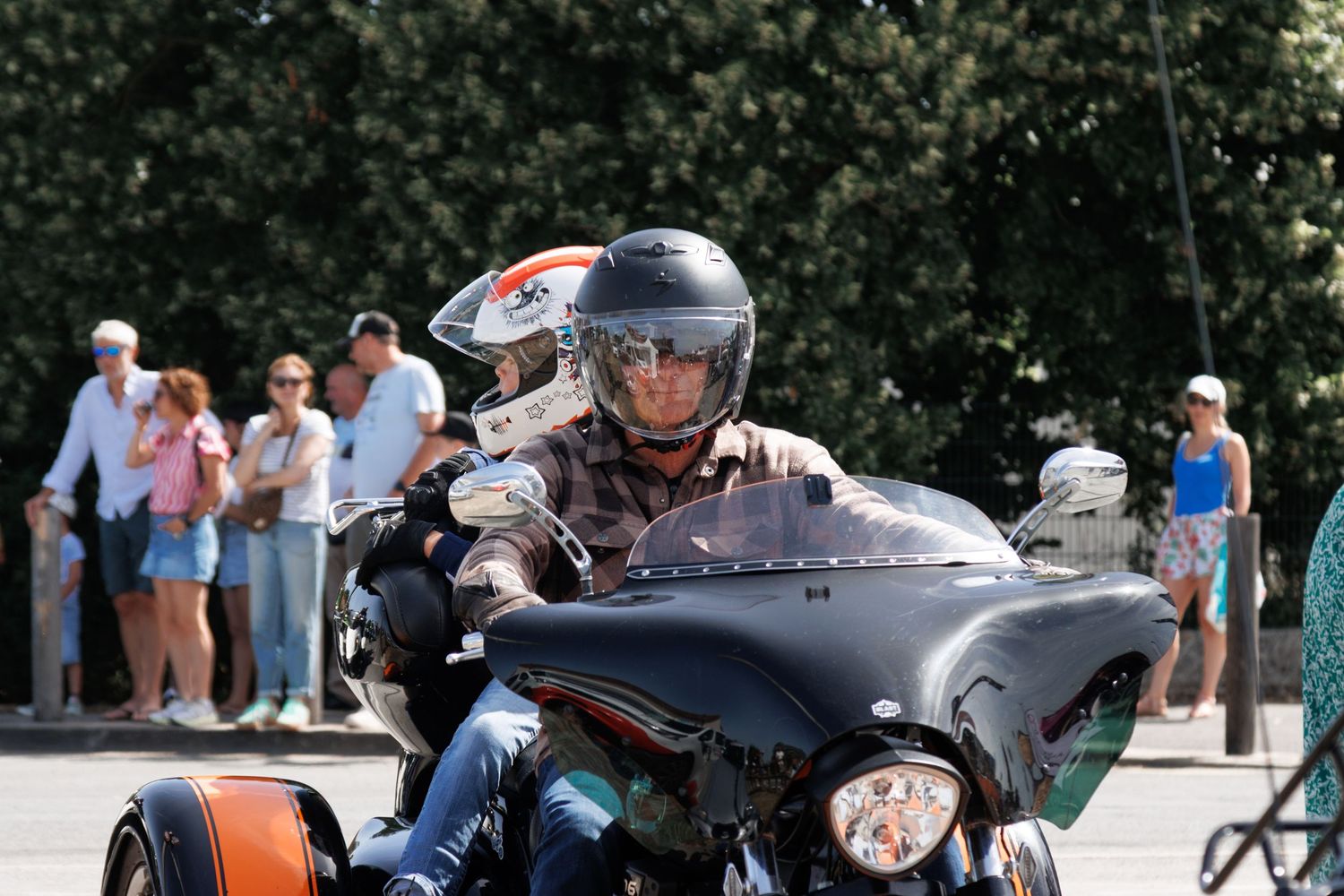 Rider in helmet on a Harley-Davidson trike motorcycle at an outdoor event with spectators in background.