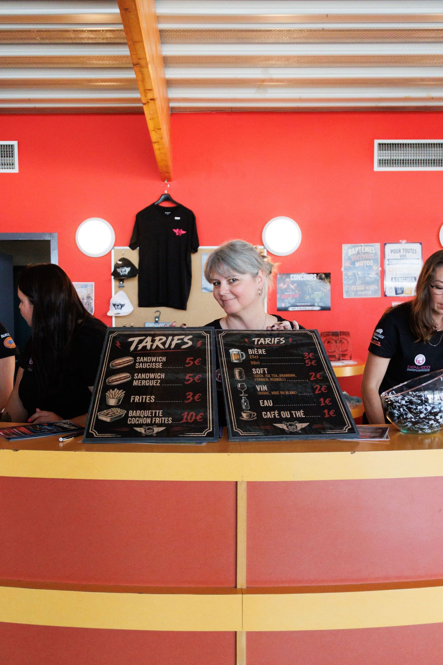 Woman smiling behind a fast food counter with French menu boards showing tarifs in a red restaurant.