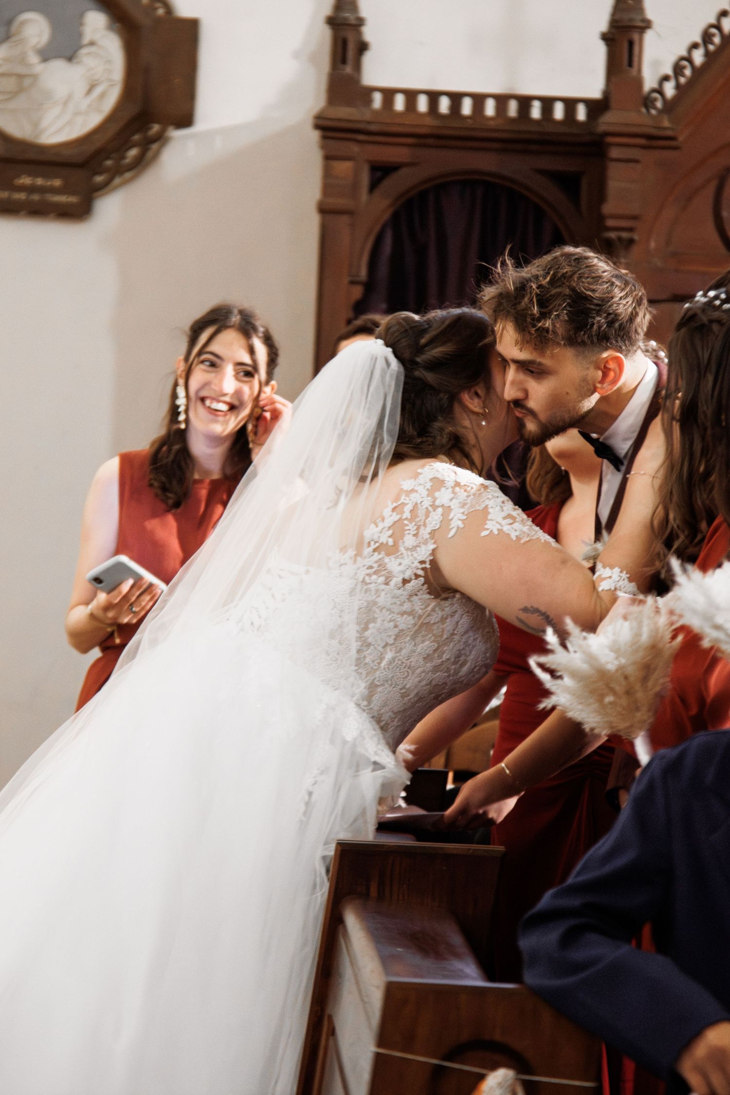 Bride and groom share a kiss at the altar during a church wedding ceremony, with a smiling bridesmaid nearby.