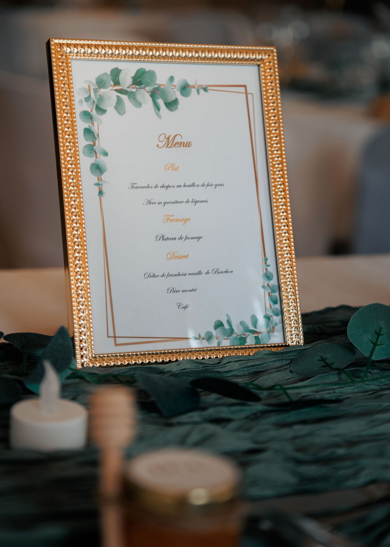 Gold framed wedding menu card with eucalyptus design on dark green table runner.