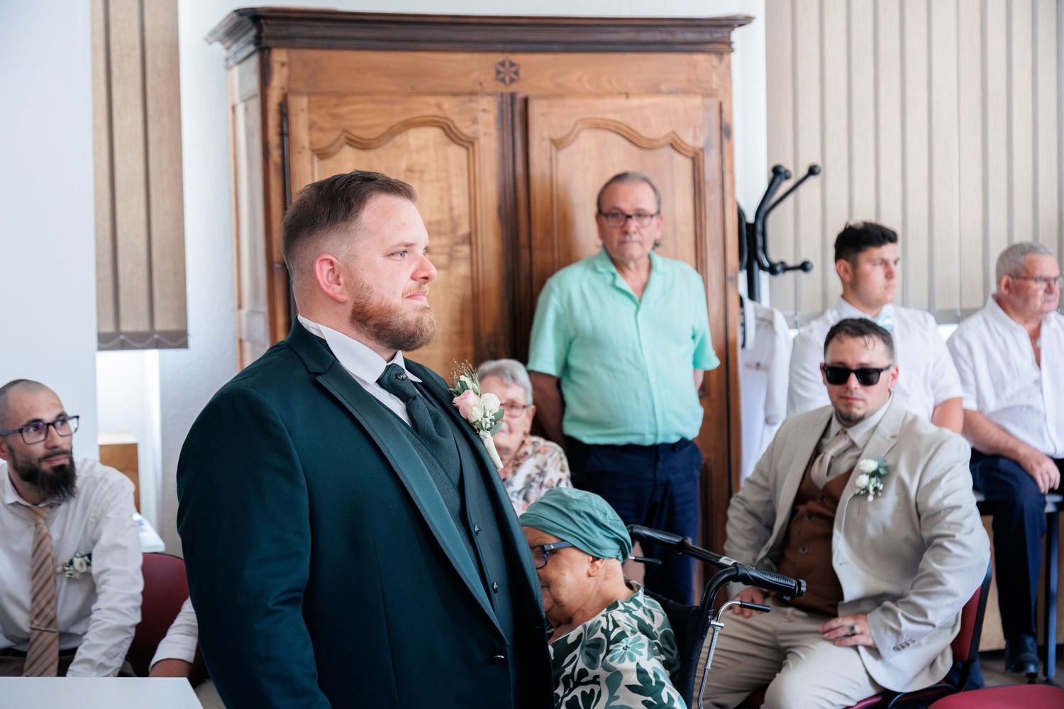 Groom in dark green suit standing at altar inside wedding ceremony hall with seated guests.