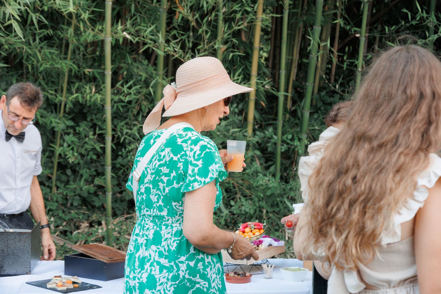 Woman in green floral dress and sun hat enjoying drinks at an outdoor wedding reception.