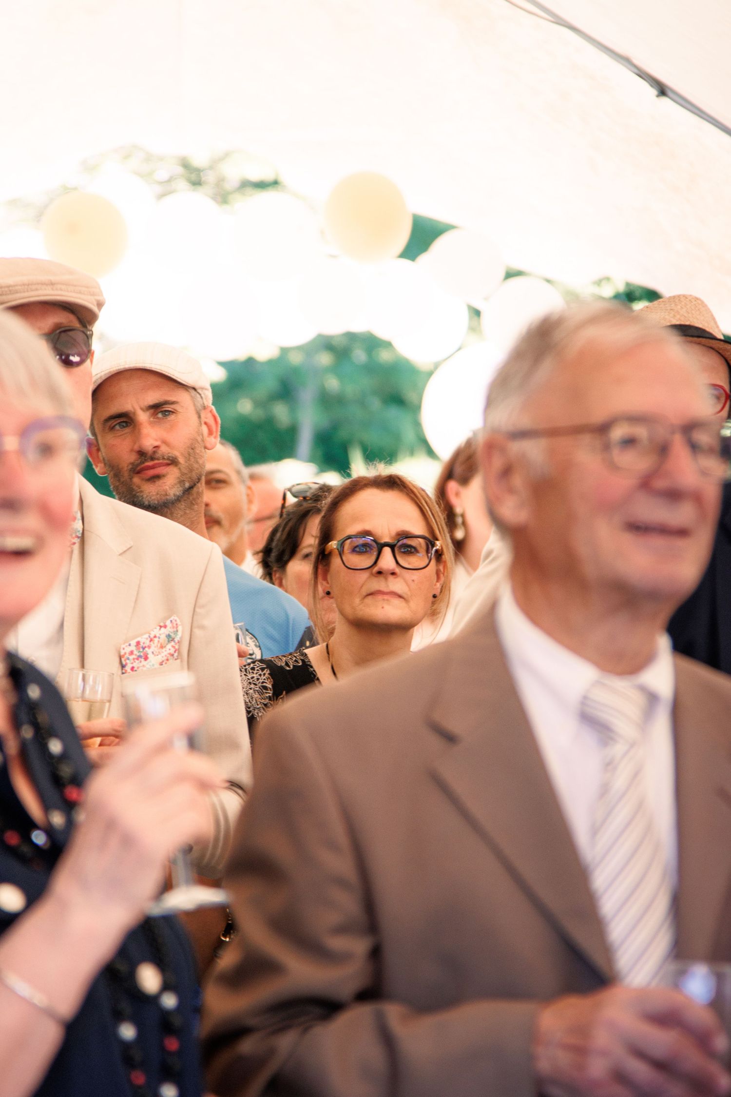 Wedding guests mingle under a white marquee tent, holding champagne glasses at an outdoor reception.