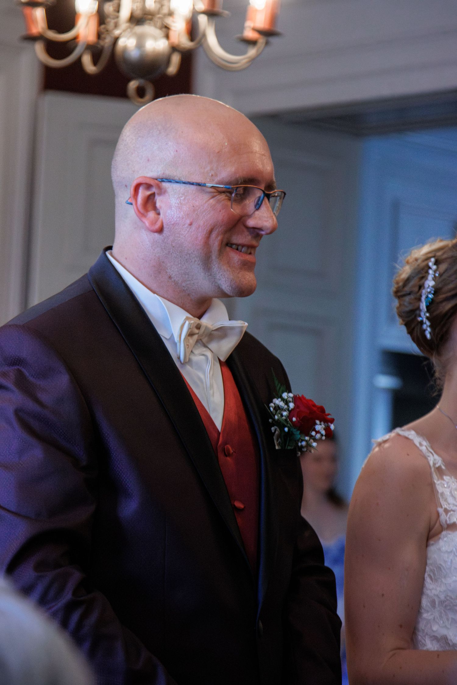 Smiling bald groom wearing glasses, red vest, and boutonniere at wedding ceremony.