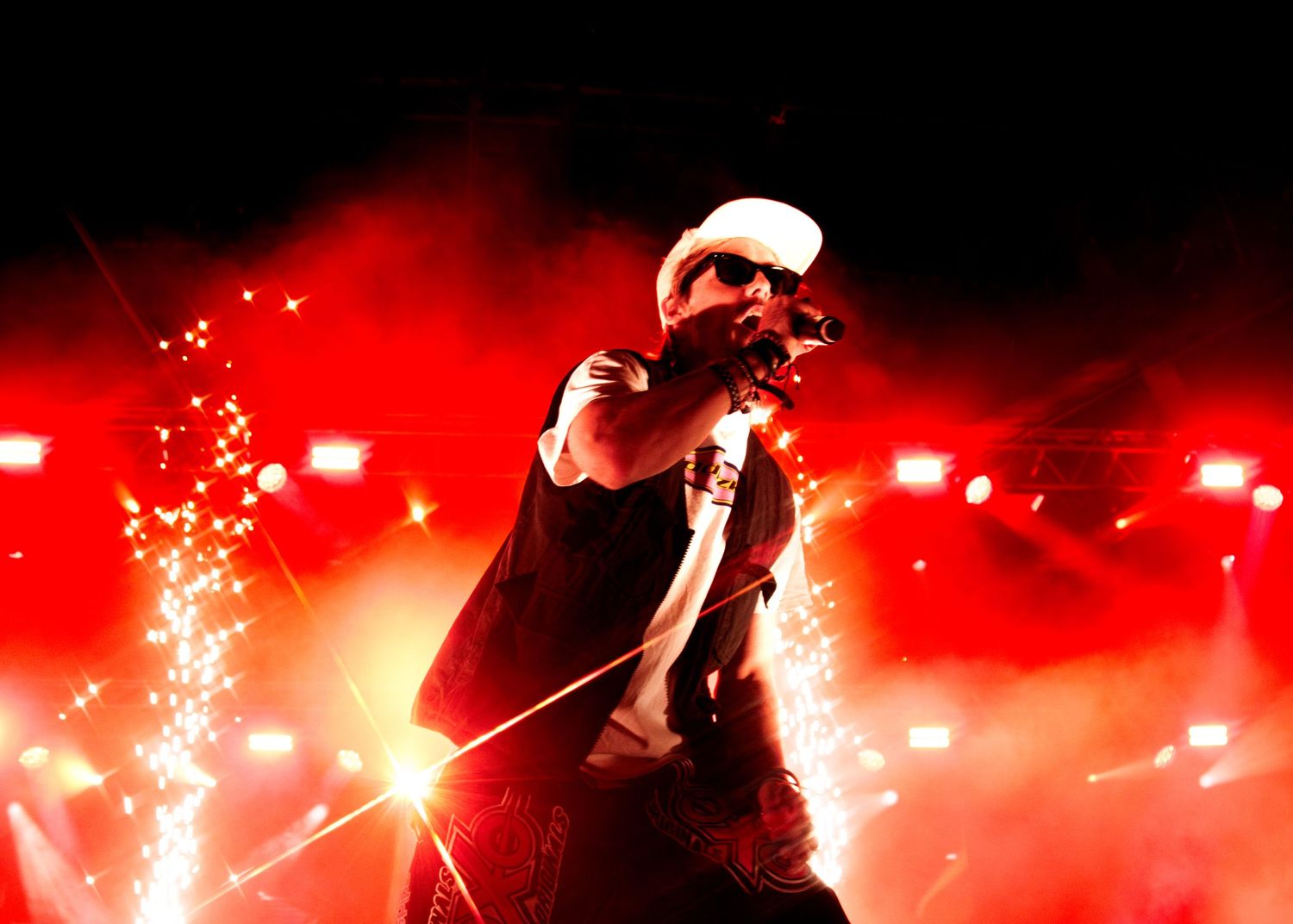 A performer in a white cap on stage surrounded by dramatic red lighting and sparkling pyrotechnic effects.
