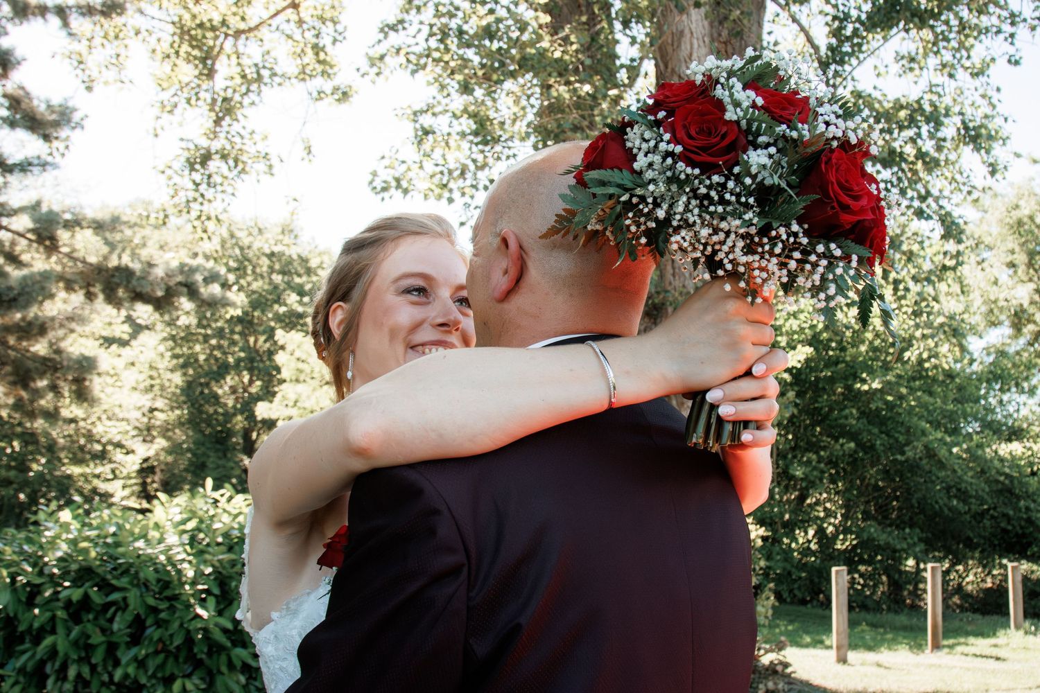 Bride in white lace gown embraces groom tenderly while holding a red roses bouquet in a garden setting.