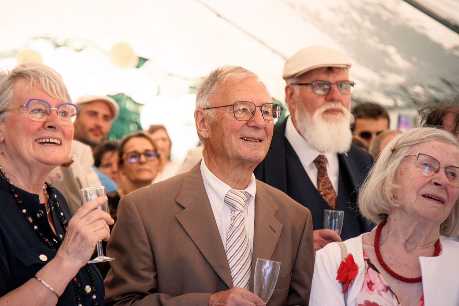 Elderly wedding guests smiling and holding champagne glasses under white tent at outdoor wedding reception.