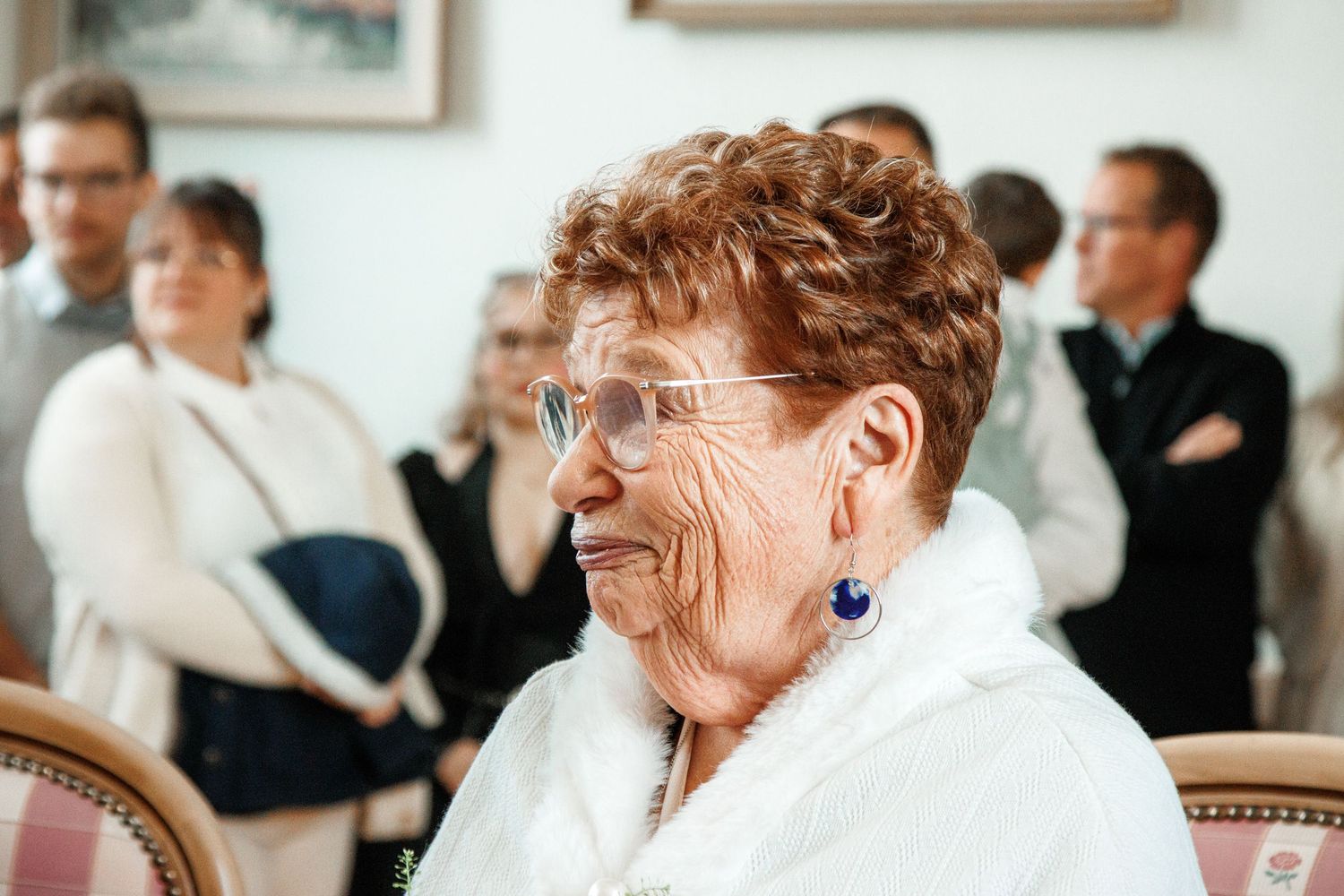Elderly woman with red curly hair and blue earrings smiling at an indoor gathering with guests in background.