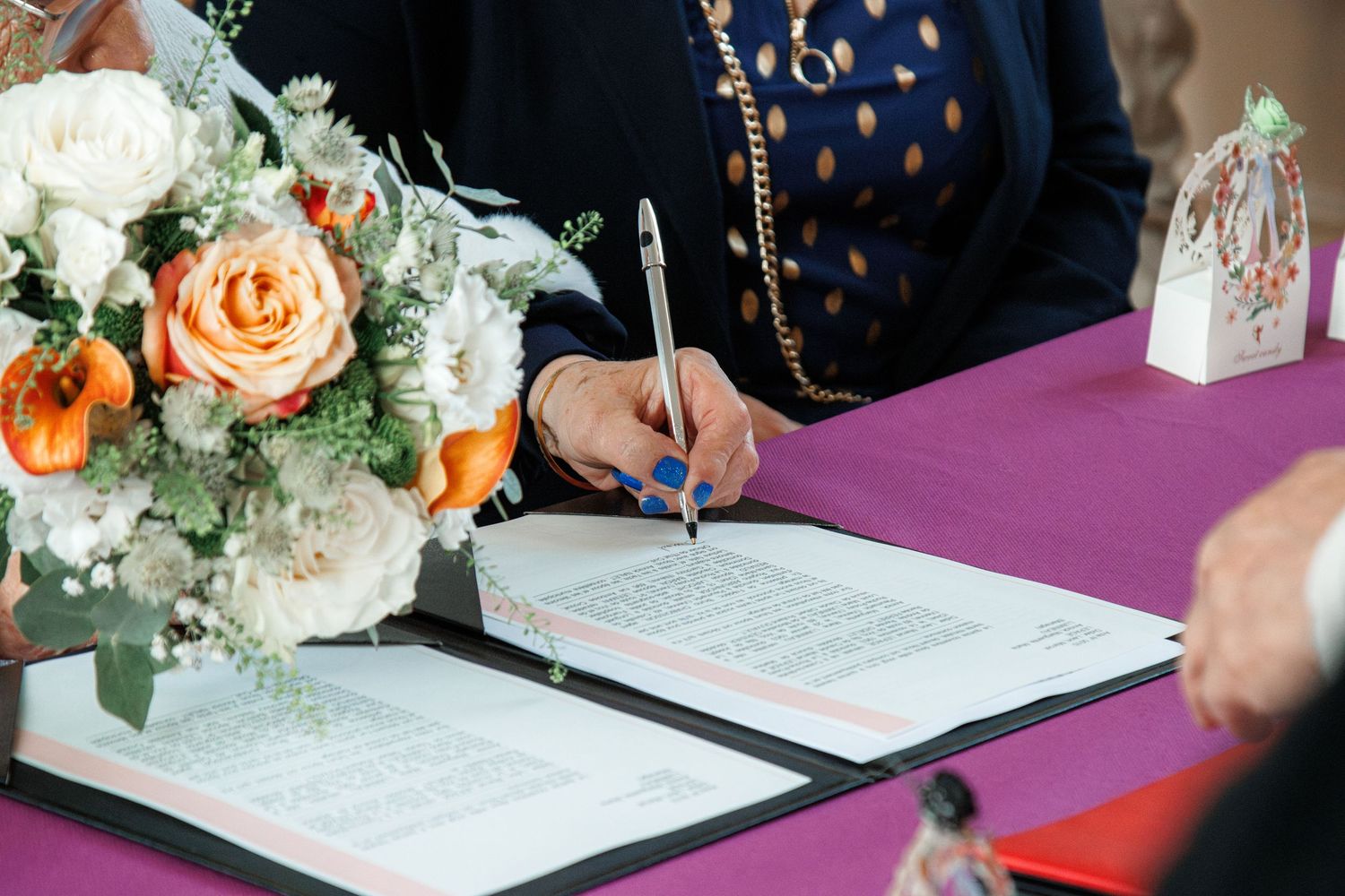 Woman signing wedding documents at a purple table with orange and white floral arrangement nearby.