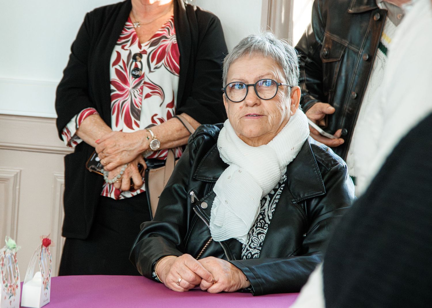 Elderly woman in black leather jacket and white scarf sitting at a table during an indoor event.