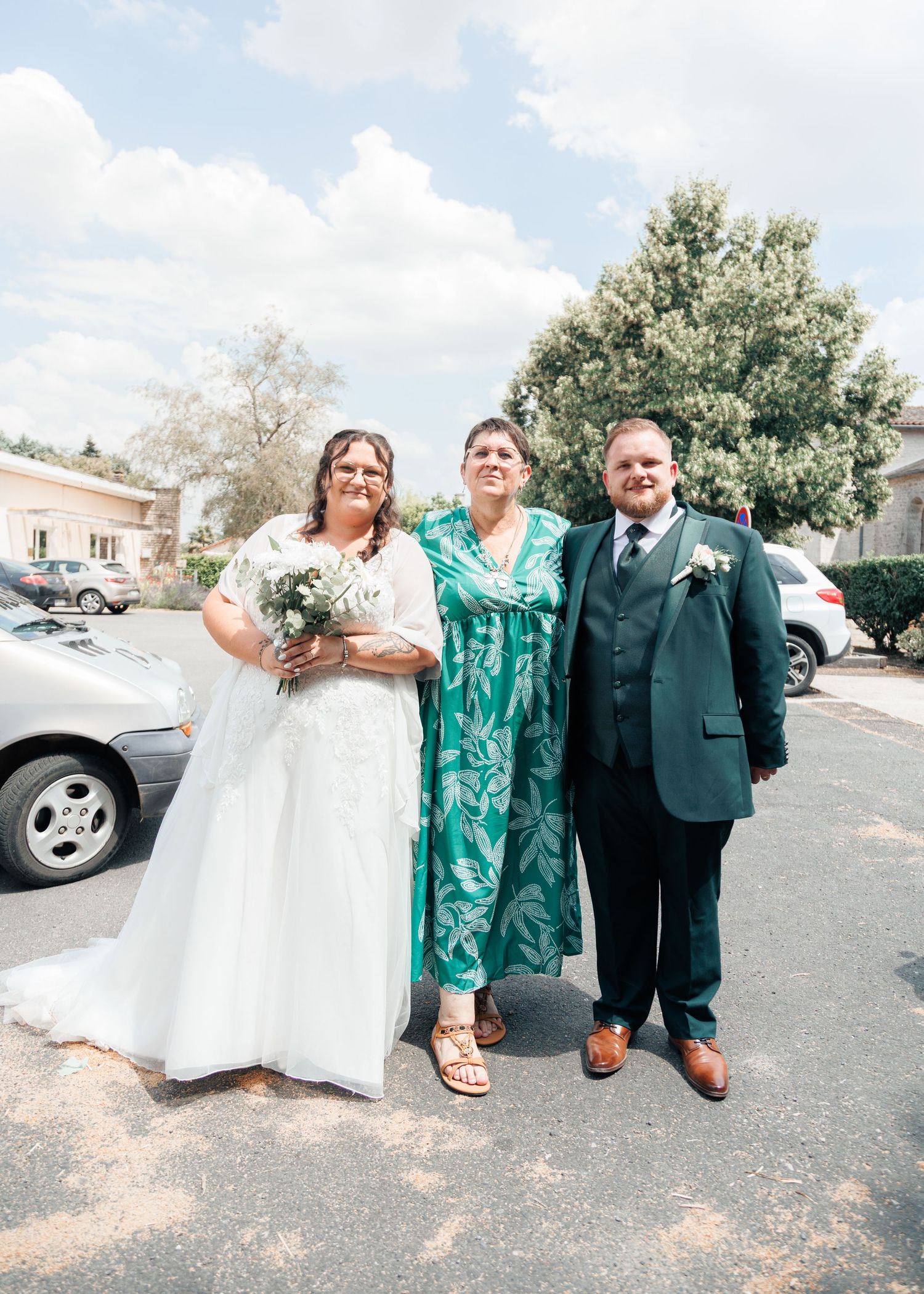 Bride in white gown and groom in green suit pose with guest in tropical dress outdoors.