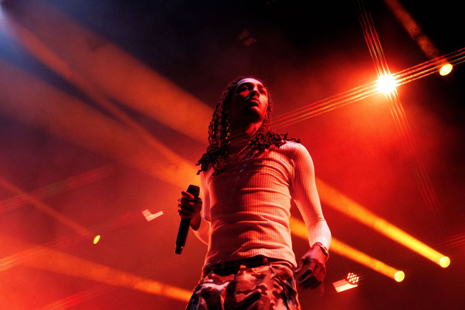 Rapper performing on stage under dramatic red and yellow stage lights, holding microphone with dreadlocks.