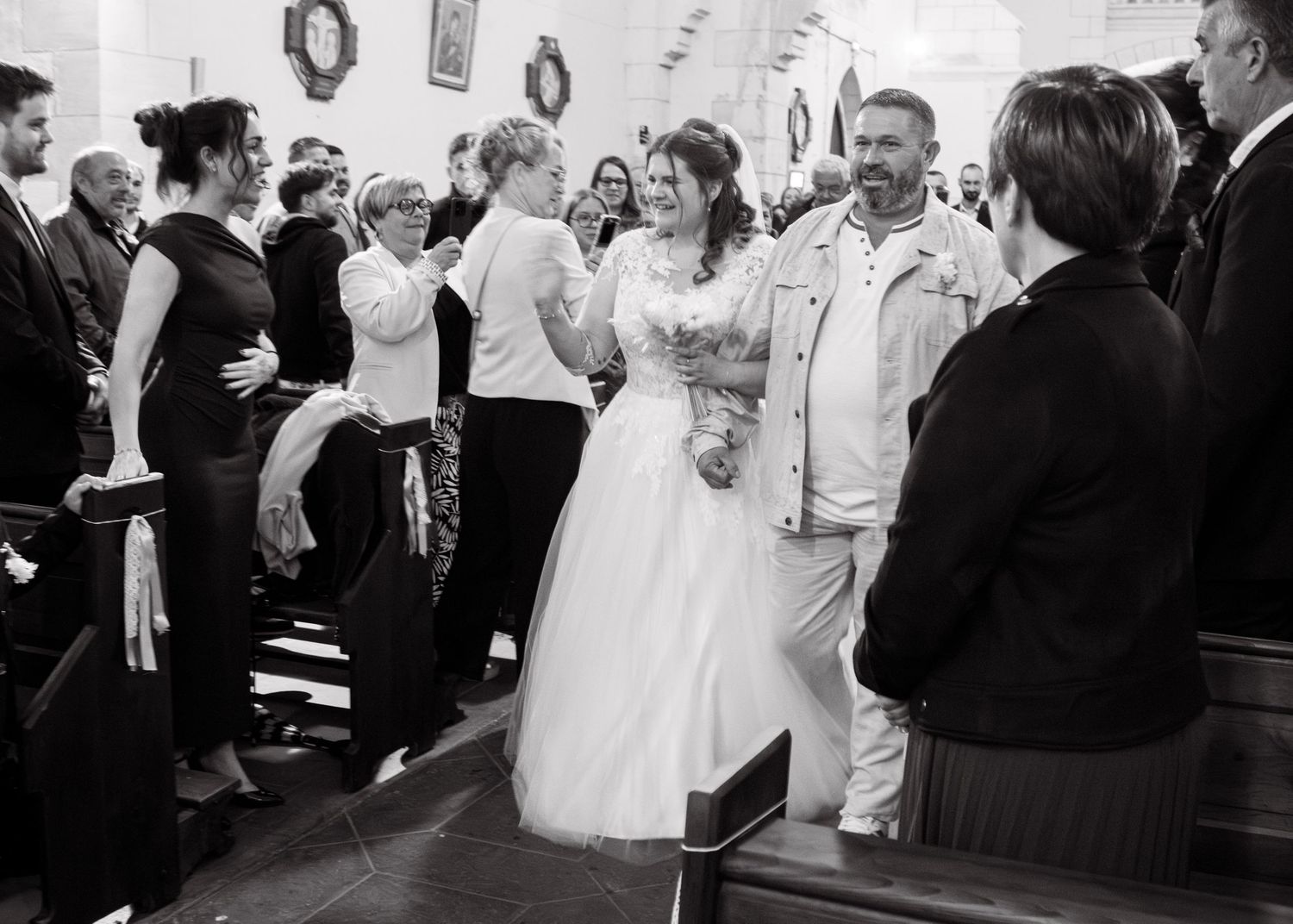 Black and white wedding photo of bride in white gown walking down church aisle with guests celebrating.