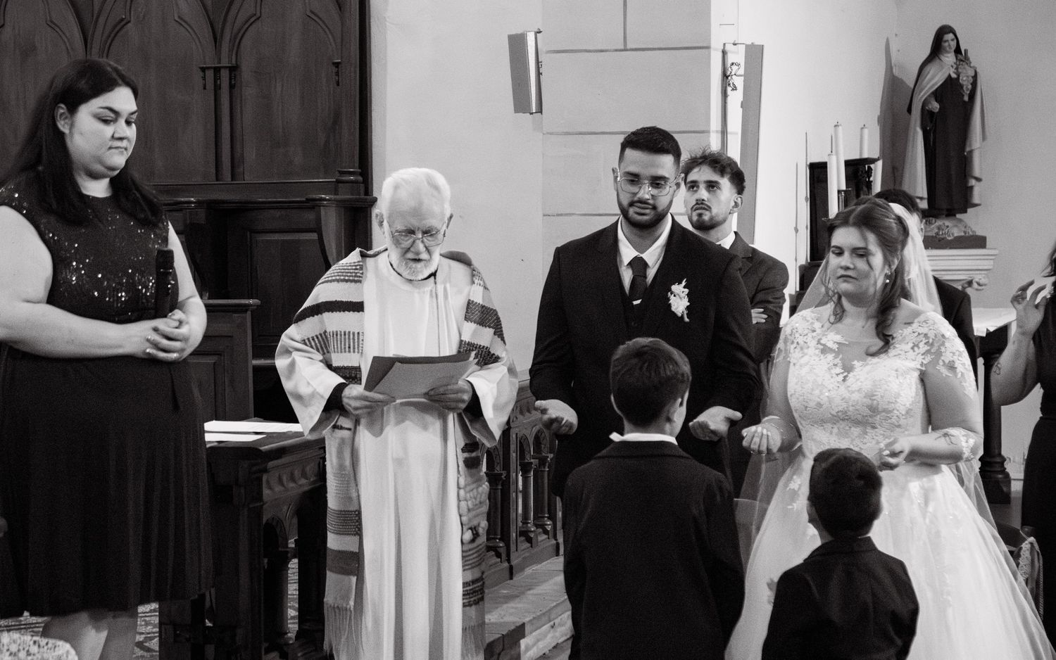 Black and white wedding ceremony photo with bride, groom, officiant, and guests in a church.