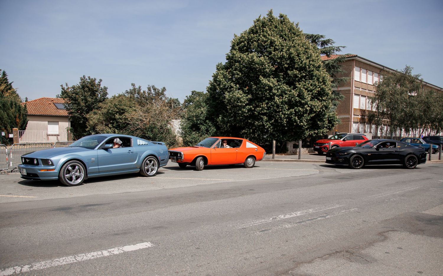 Three classic muscle cars including a blue Ford Mustang and orange muscle car parked on a sunny street near a brick building.