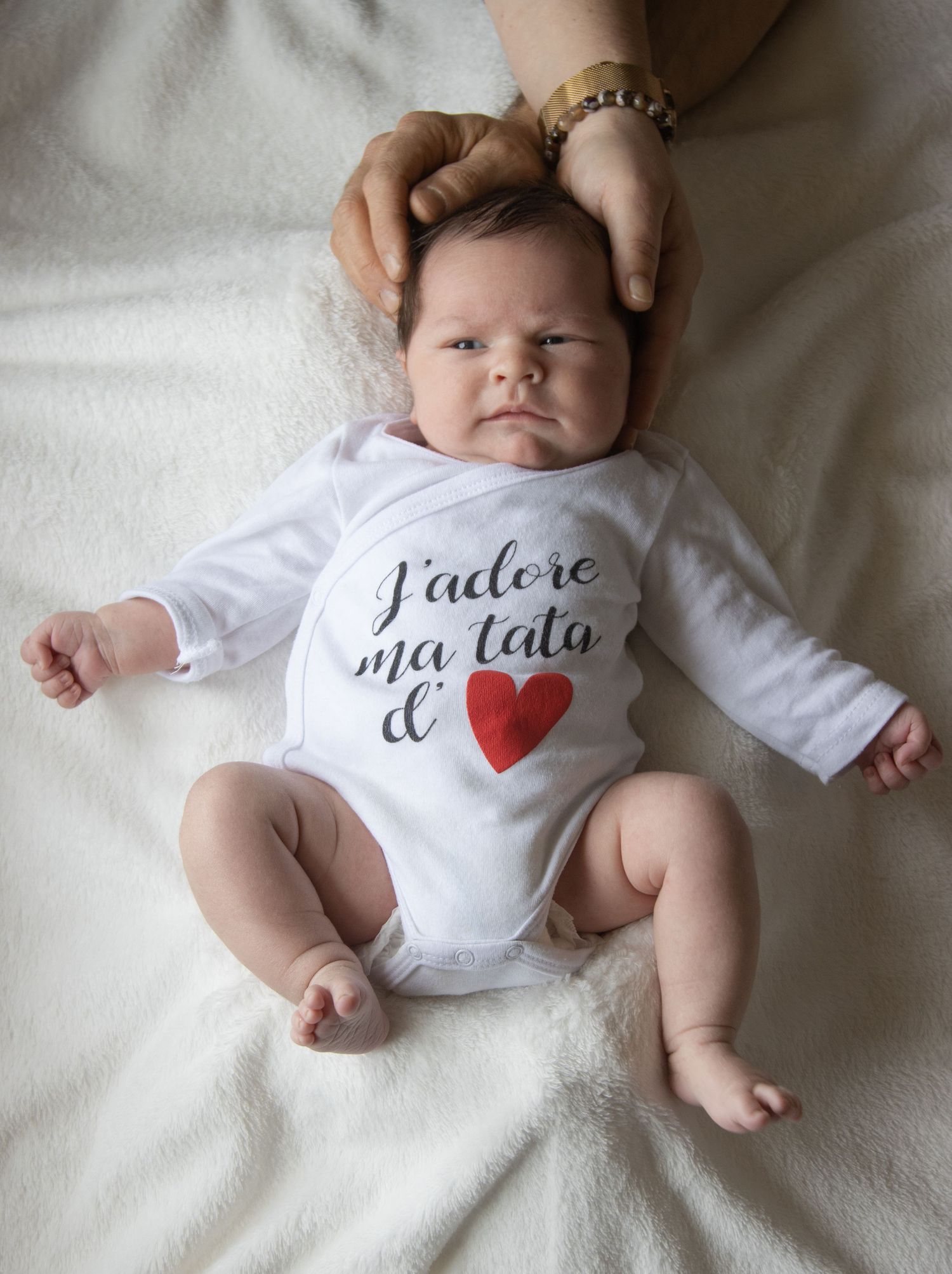A baby wearing a white onesie with French text and heart design lies on cream colored fabric.