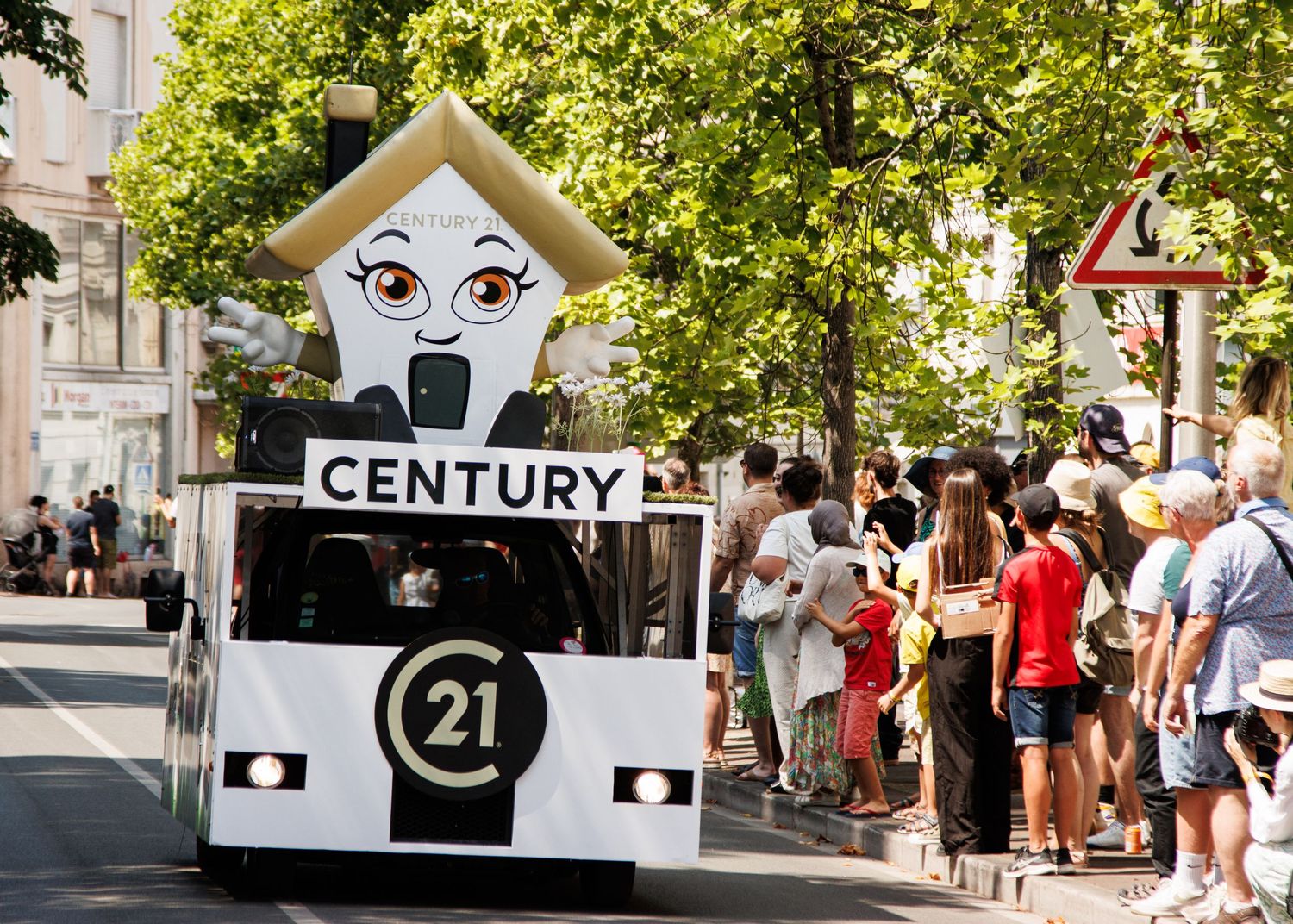 Century 21 themed parade float with cartoon house design rolls through crowded street festival.