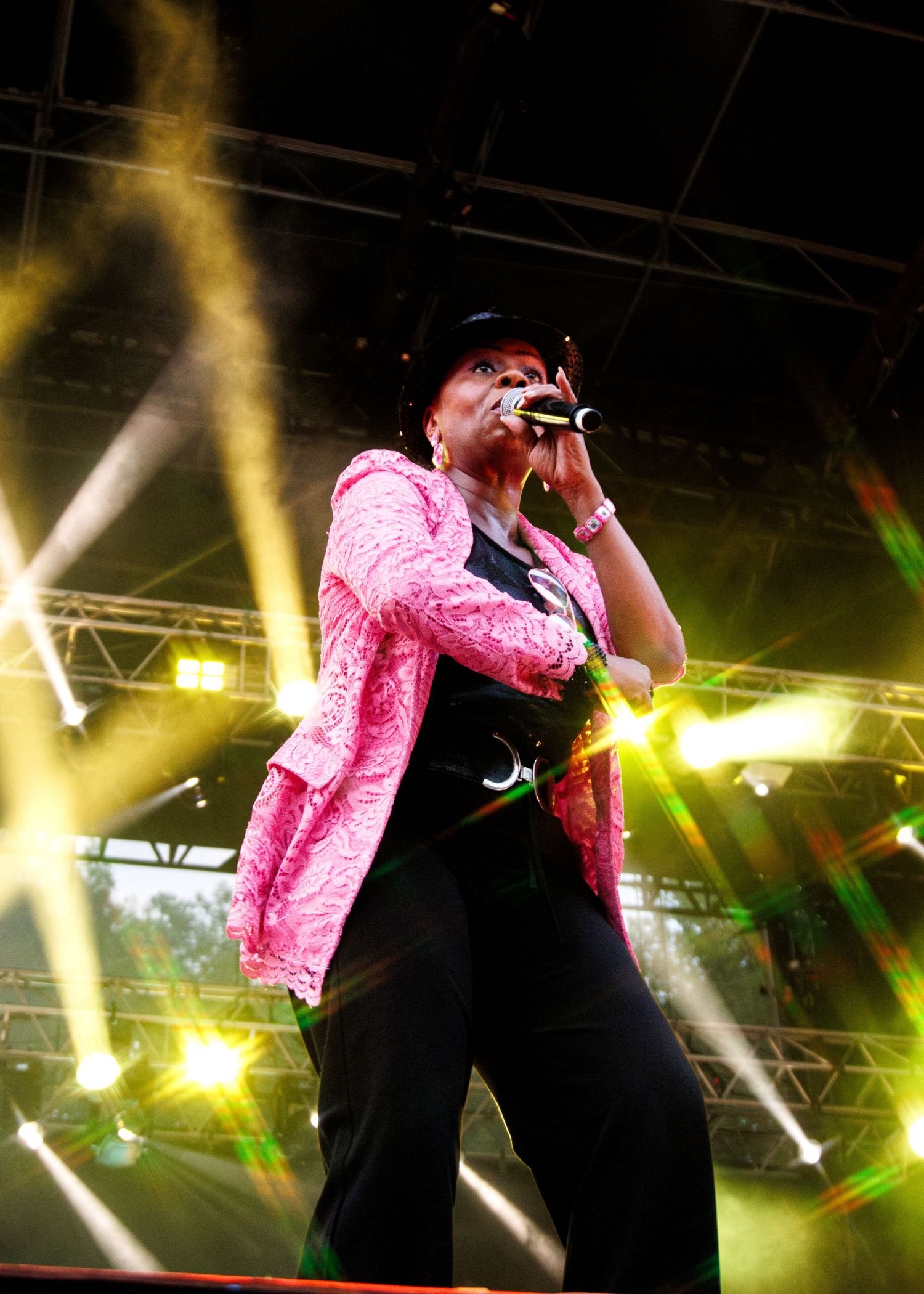 Performer in pink sequined jacket and black pants sings on stage with dramatic yellow lighting and beams in background.