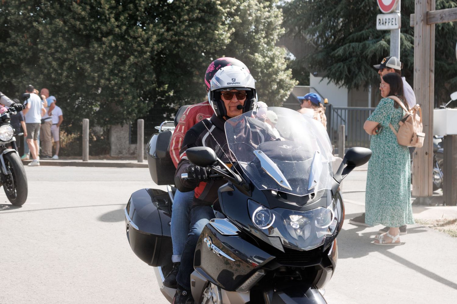 Two people riding a large touring motorcycle on a sunny street, with pedestrians and greenery visible in the background.