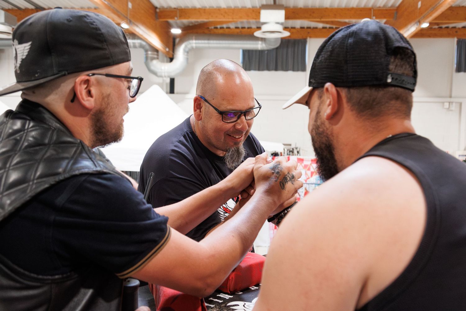 Three men collaborating on a tattoo application at an indoor tattoo convention event.