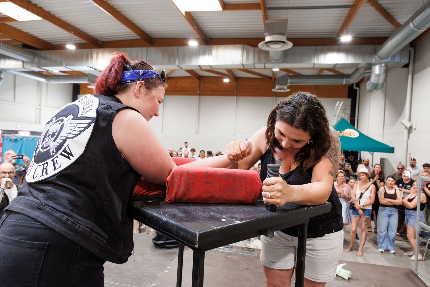 Two women competing in an arm wrestling match at an indoor event with a crowd watching in the background.