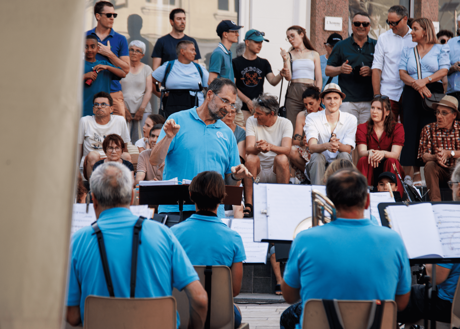 A band conductor directs musicians during an outdoor performance with audience members watching.