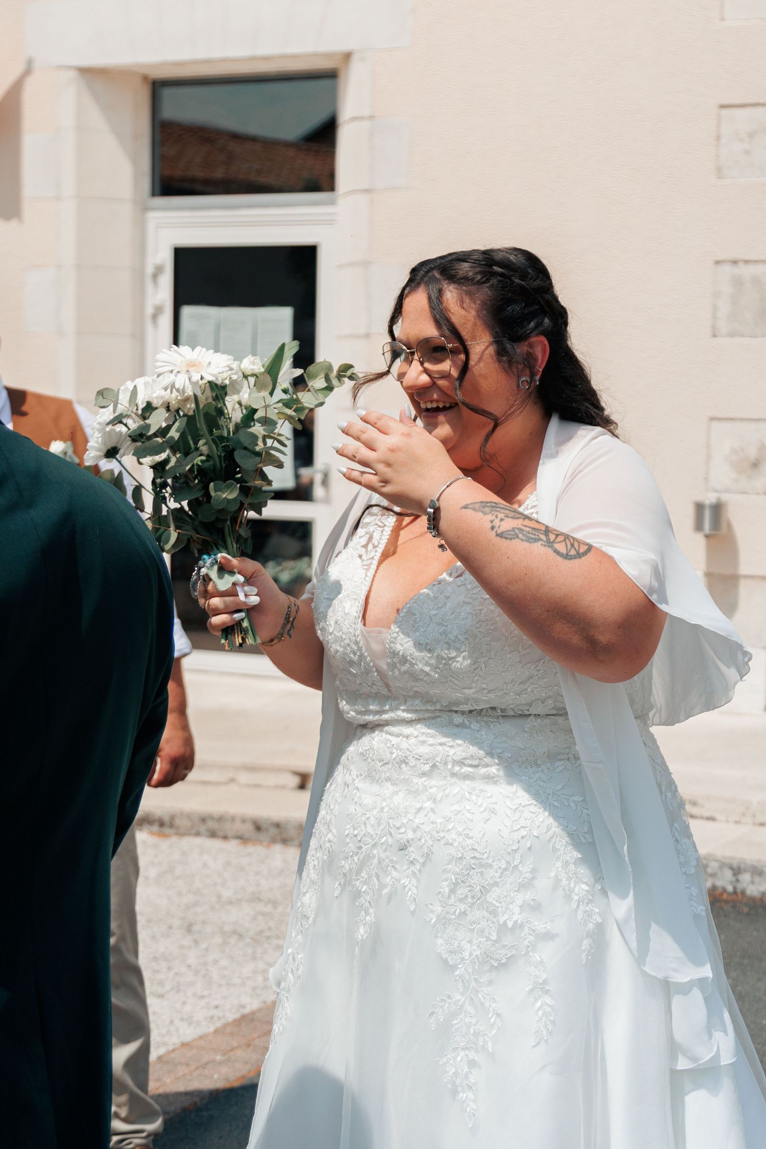 Emotional bride in white lace dress holding white bouquet outside church on wedding day.