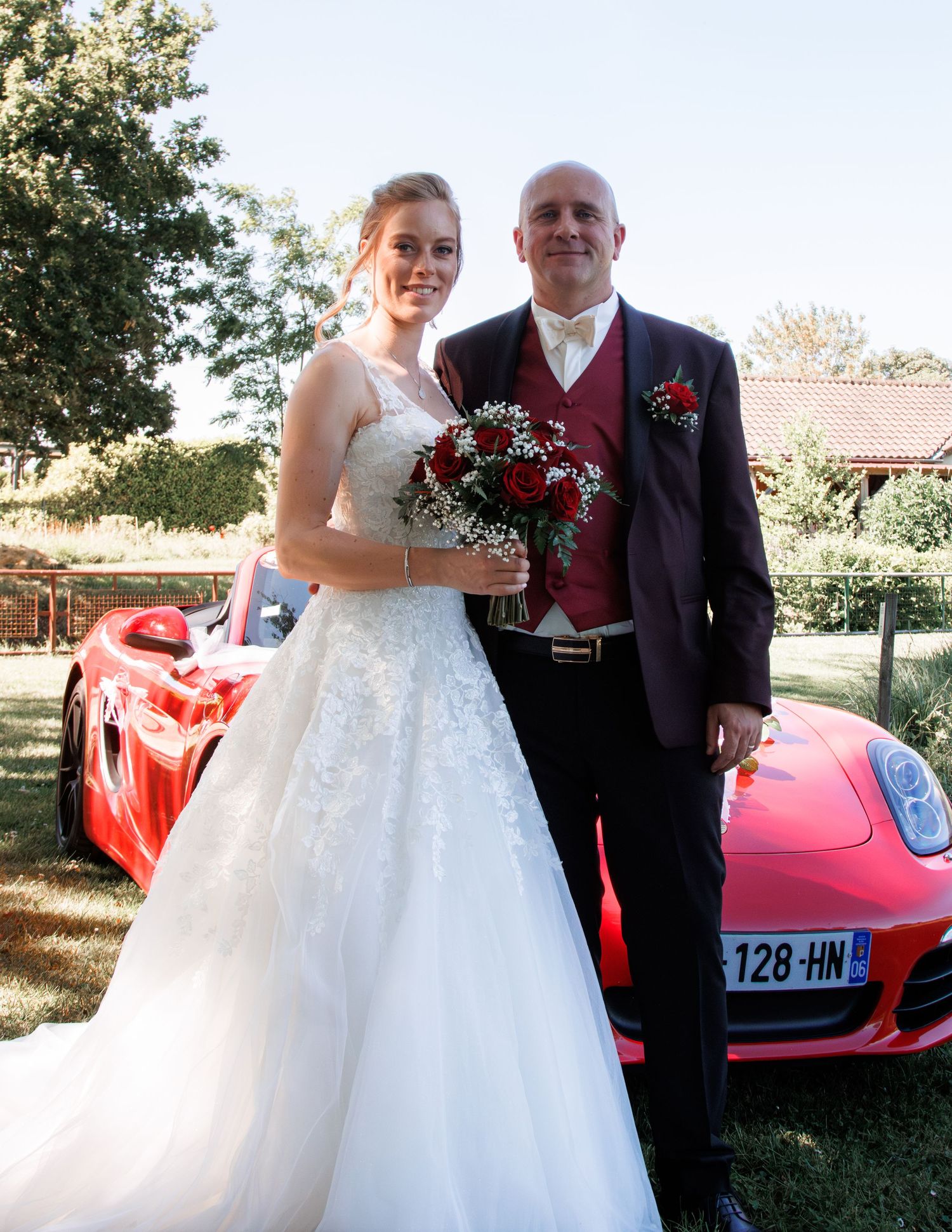 Bride in white gown holding red bouquet poses with groom in dark suit beside a red Porsche on wedding day.