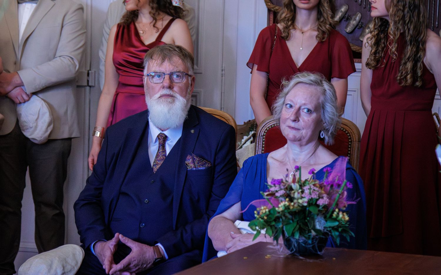 Elderly couple in formal attire seated at a table with flowers, surrounded by people in red dresses at a celebration.