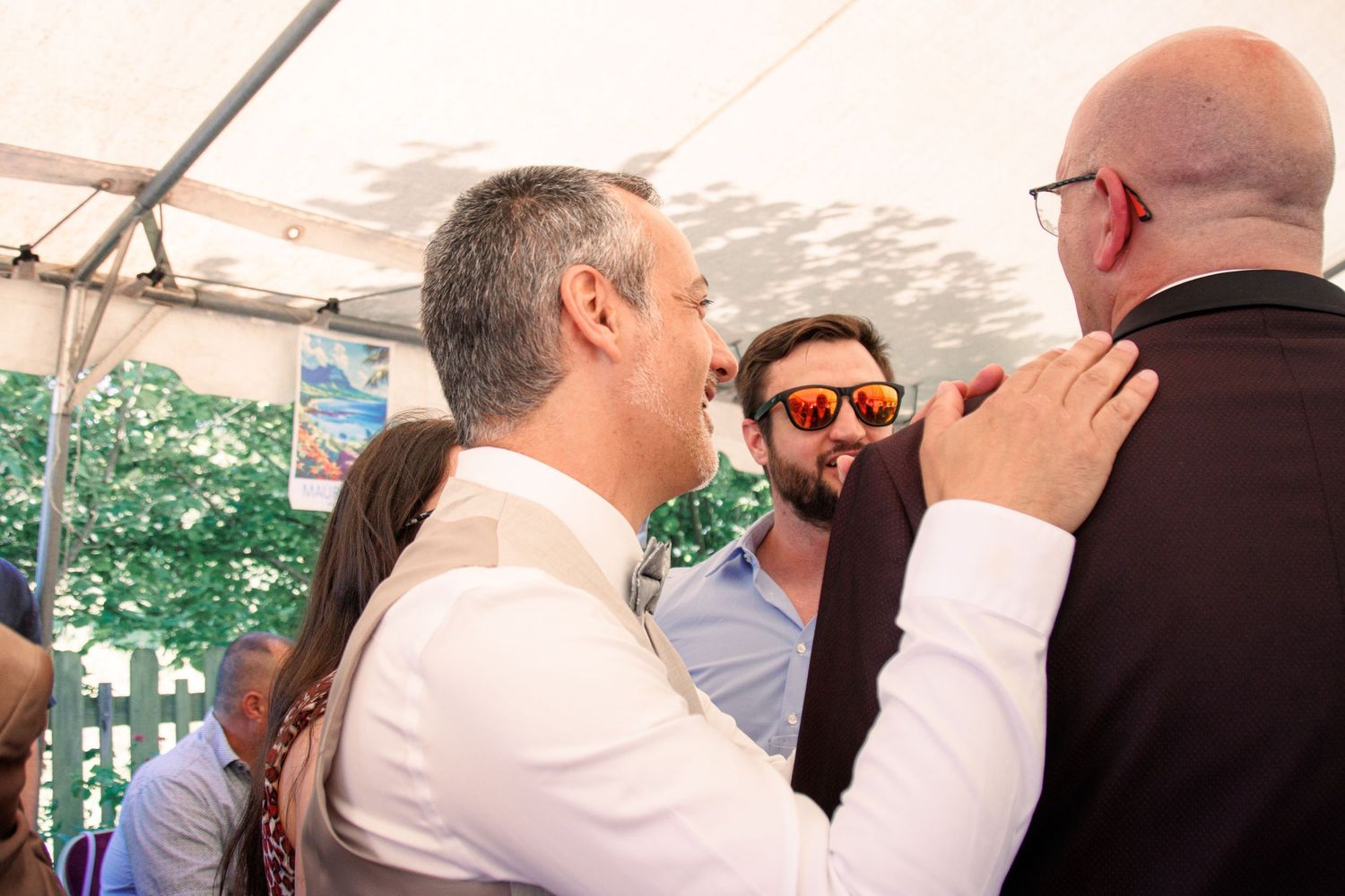 Two men in suits share a warm greeting at an outdoor wedding reception under a white tent.