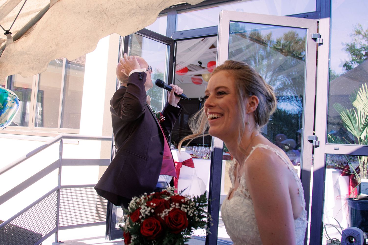Joyful bride laughing holding red bouquet while groom waves microphone at wedding reception.