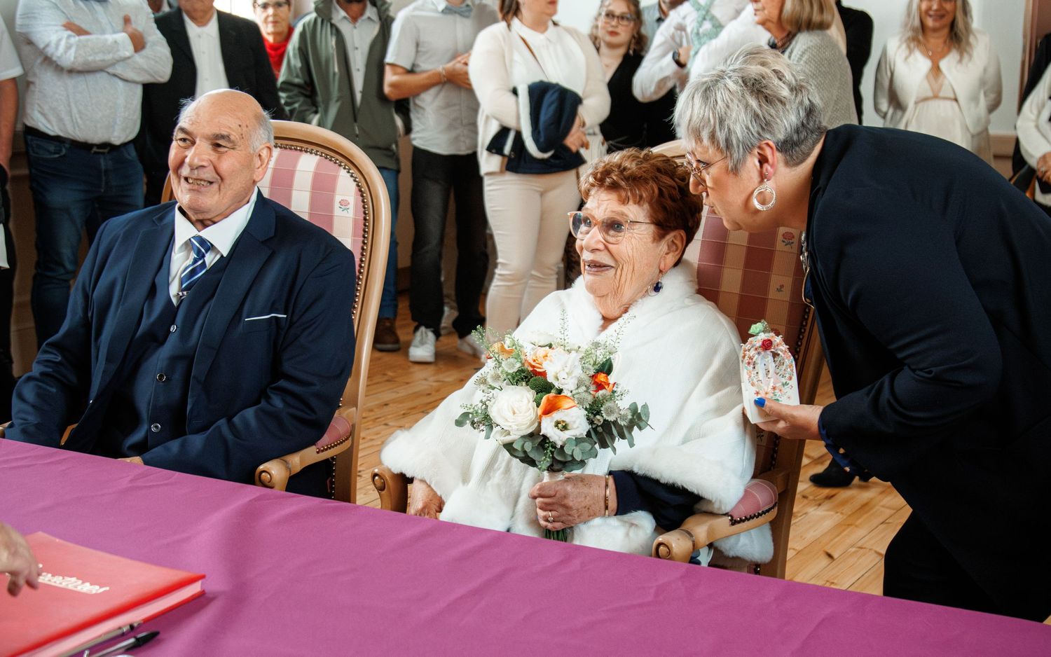 Elderly couple at civil wedding ceremony, bride holding white bouquet, guests watching in elegant hall.