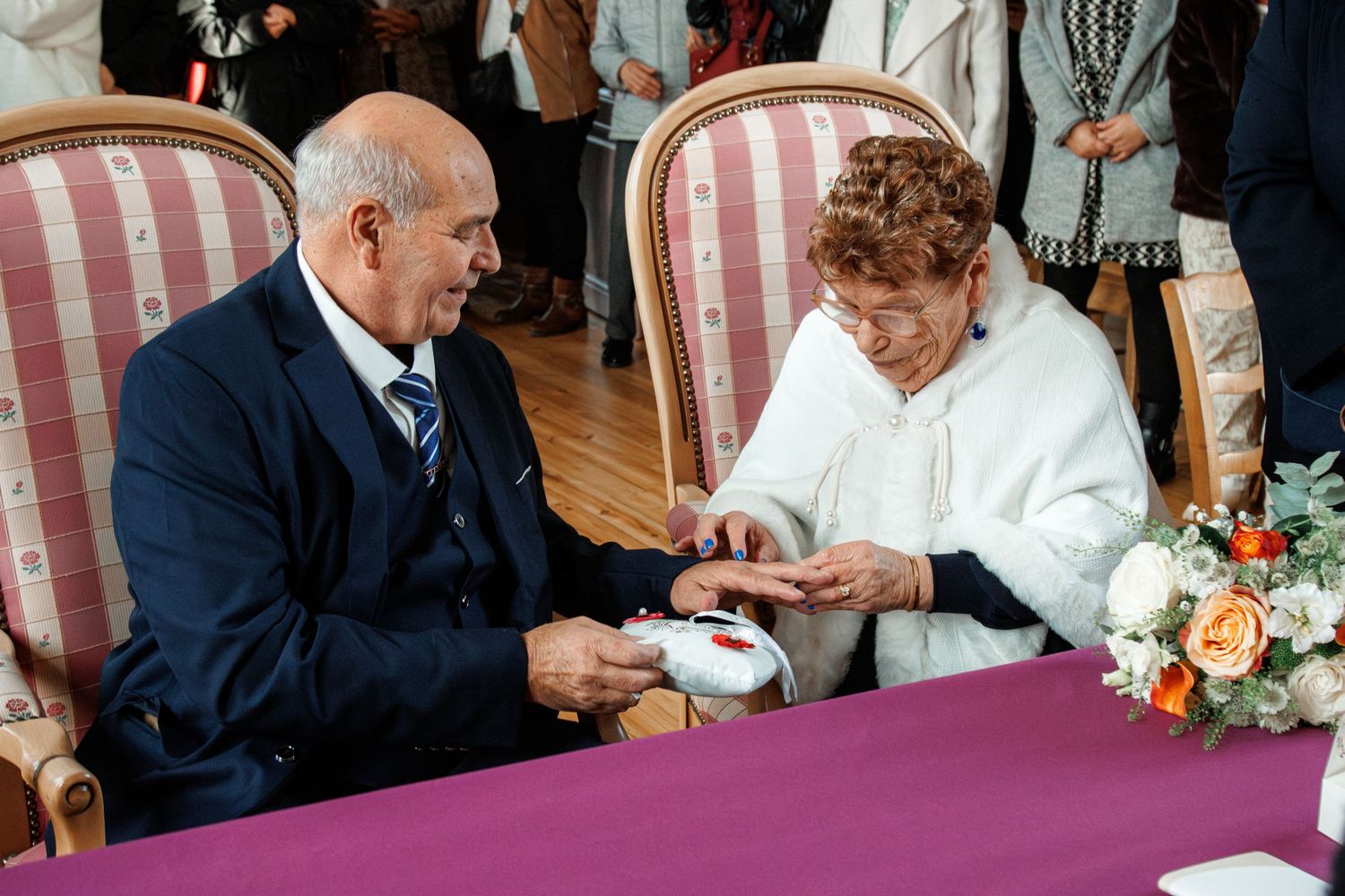 Elderly couple exchanging rings at a wedding ceremony, seated at a purple-draped table with flowers.