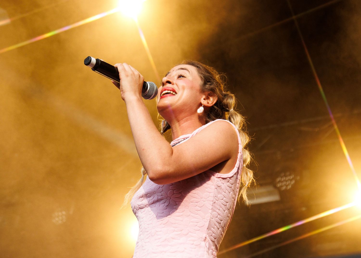 A performer sings passionately into a microphone on stage with dramatic golden lighting and lens flares in the background.