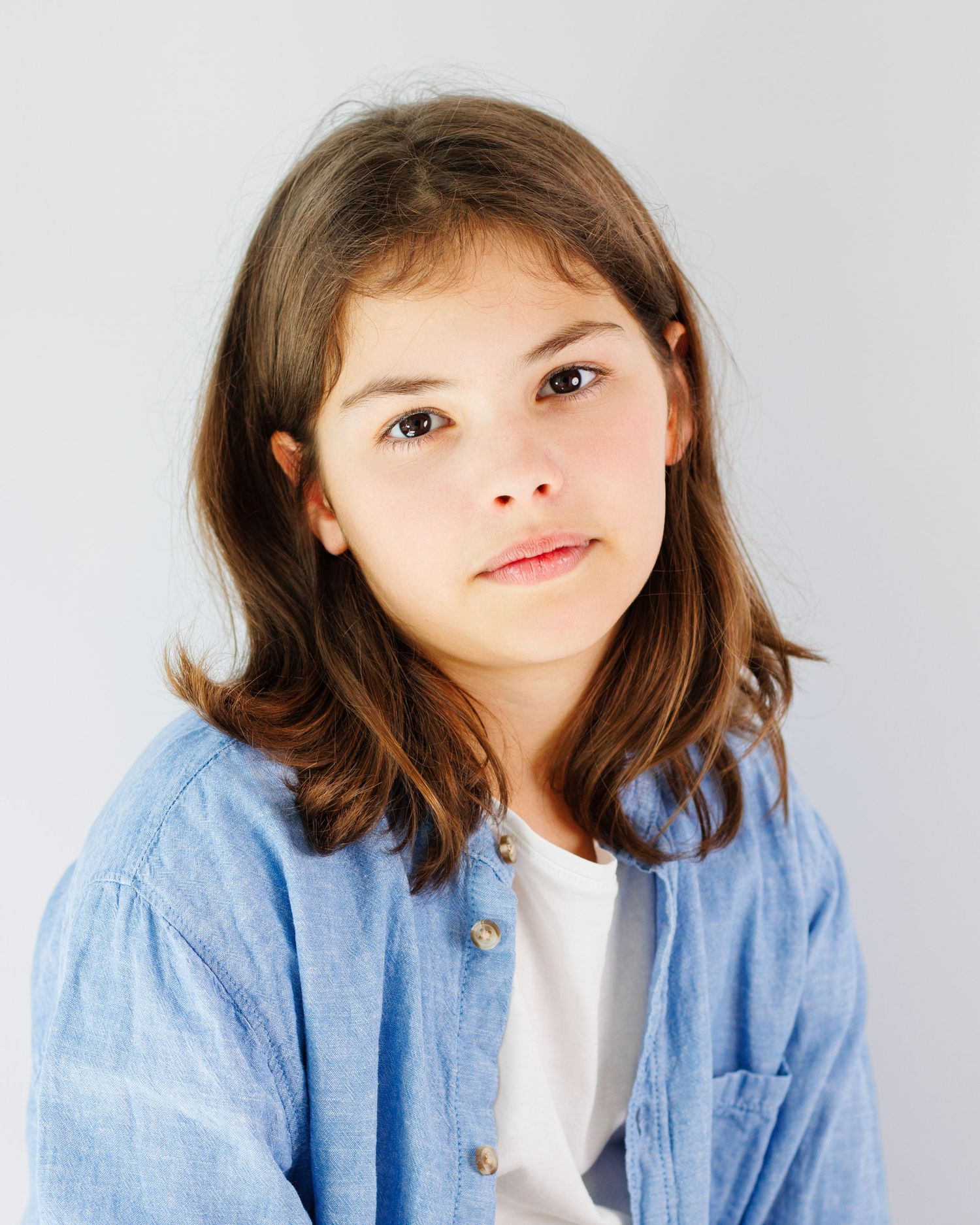 Young person wearing a light blue button-down shirt over a white top against a plain background.