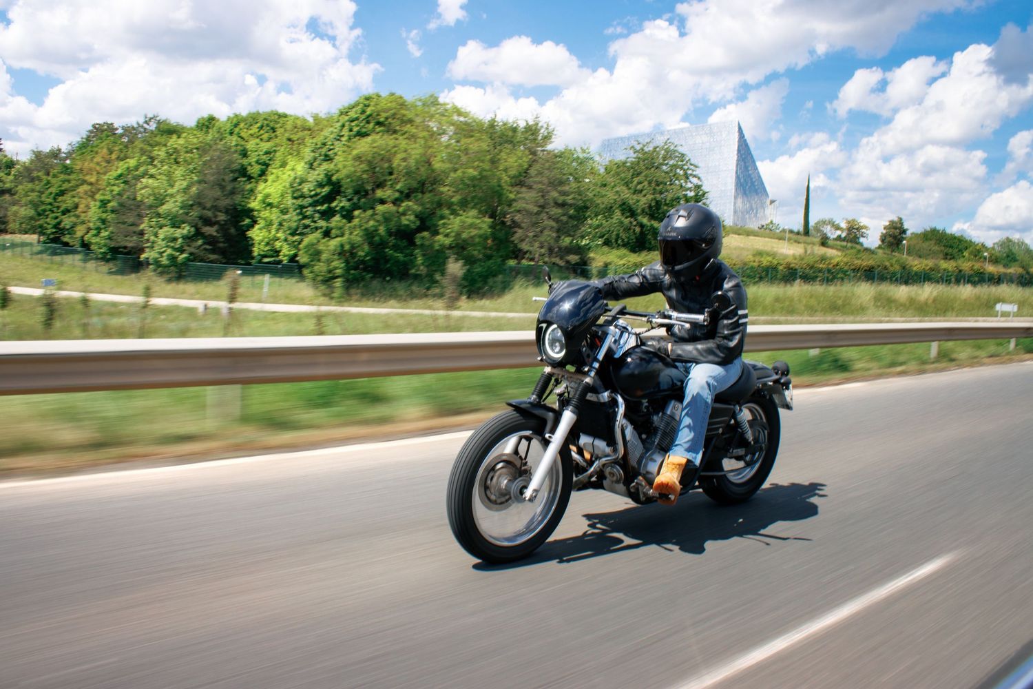 Motorcyclist riding a black motorcycle on a highway surrounded by green trees under a partly cloudy blue sky.