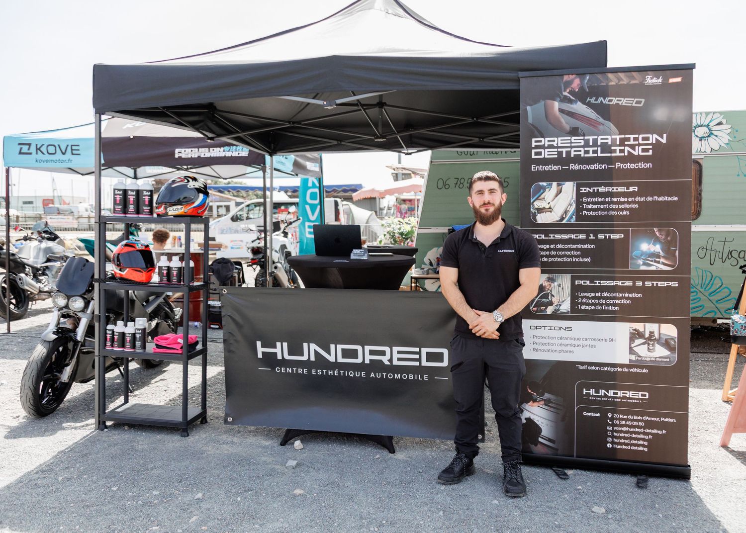 Man stands at Hundred auto detailing brand booth with products display at outdoor motorsport event.