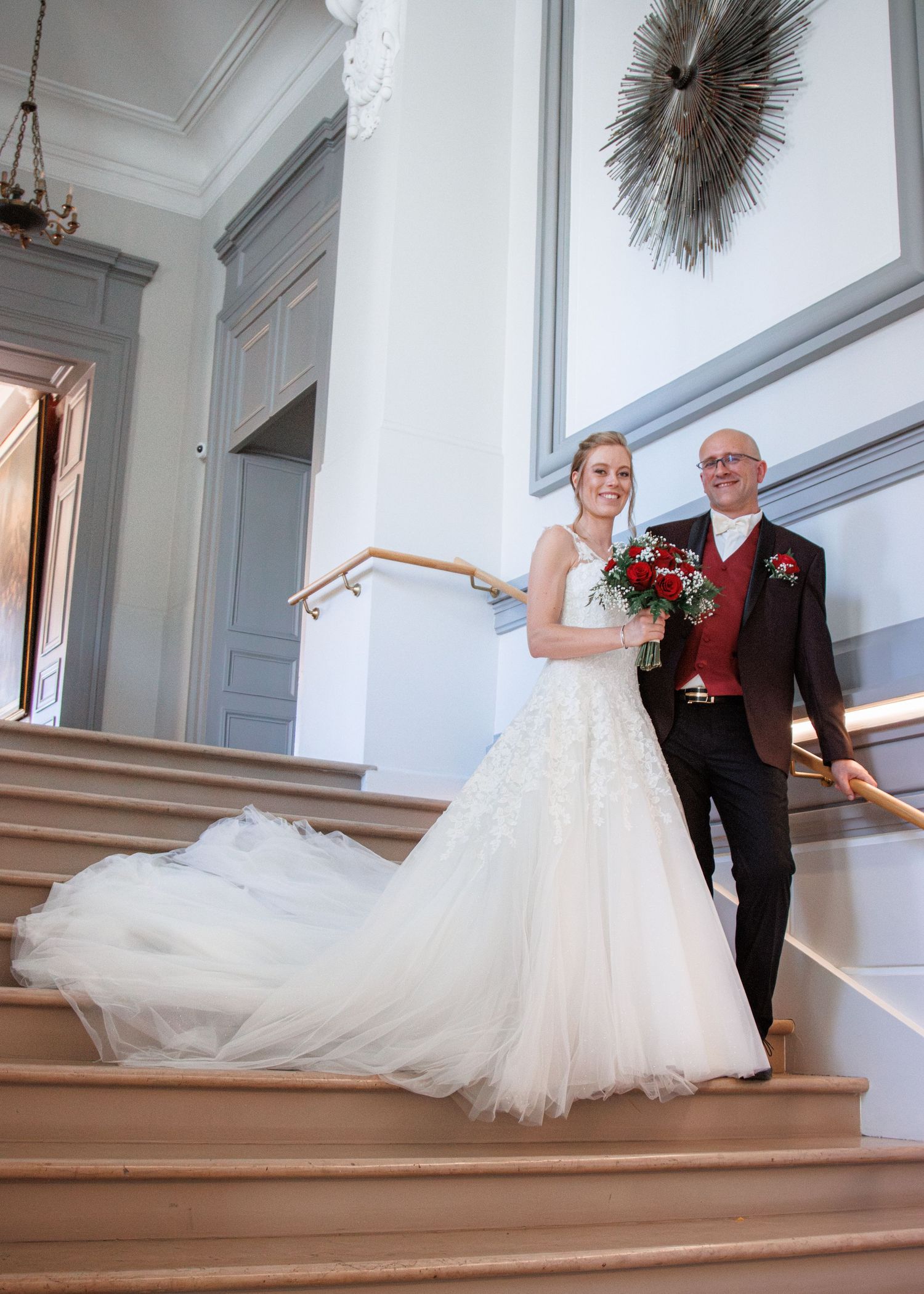 Bride in white lace gown with red bouquet poses with father in burgundy suit on elegant staircase.