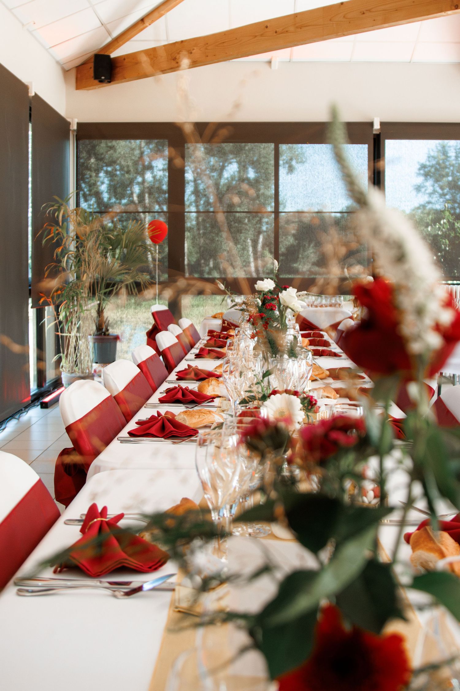 Elegant wedding reception table set with red napkins, floral centerpieces, and glassware in a sunlit venue.