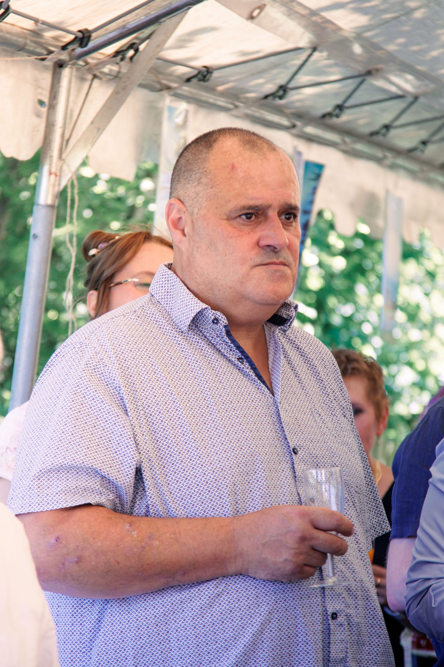 Middle-aged man in a patterned shirt holding a glass at an outdoor event under a white tent.