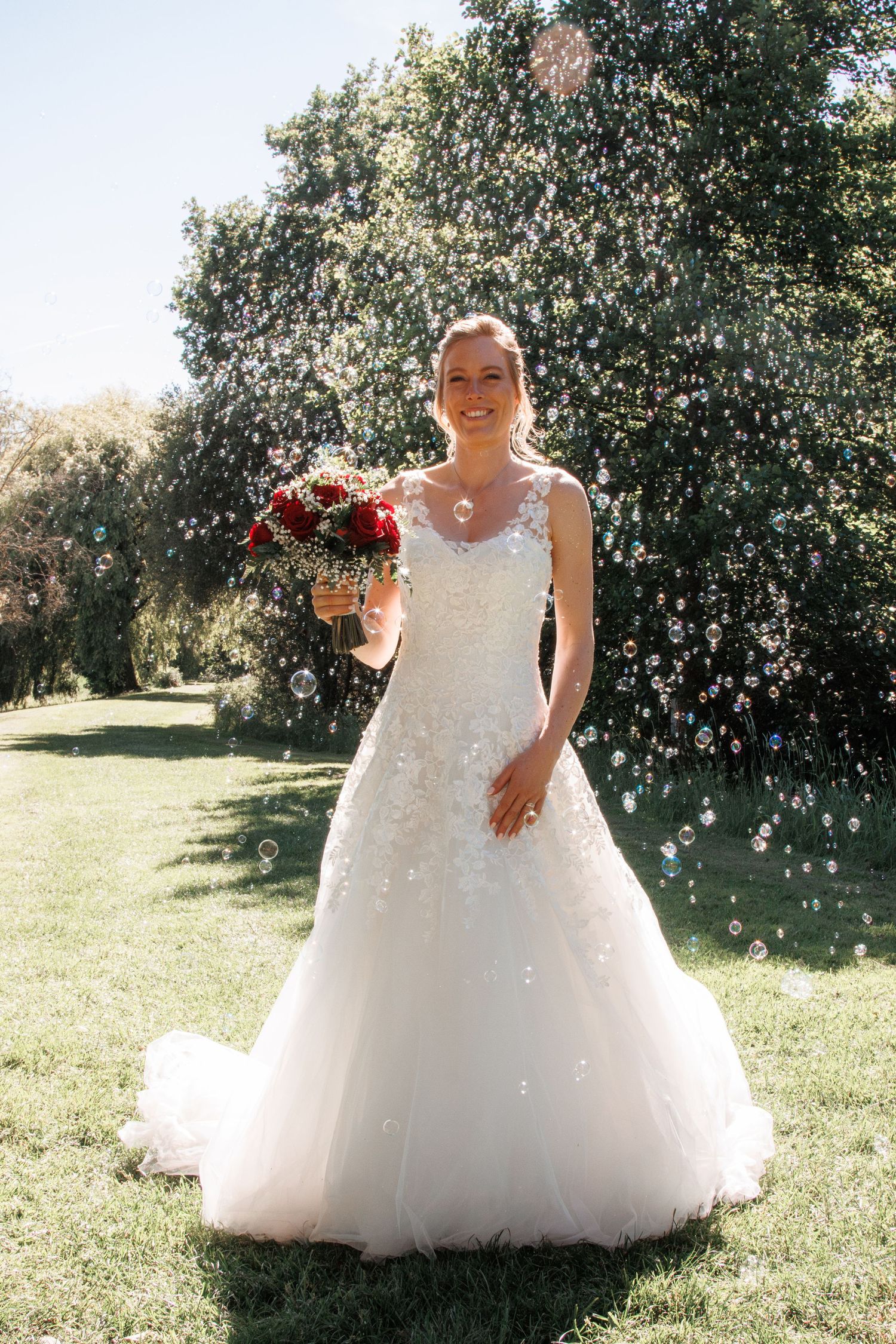 Beautiful bride in white lace gown holding red bouquet surrounded by floating bubbles outdoors.