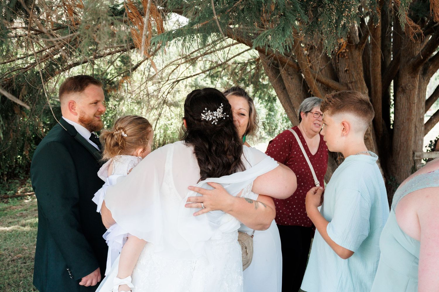 Bride in white gown receiving warm hugs from family and friends during outdoor wedding ceremony.