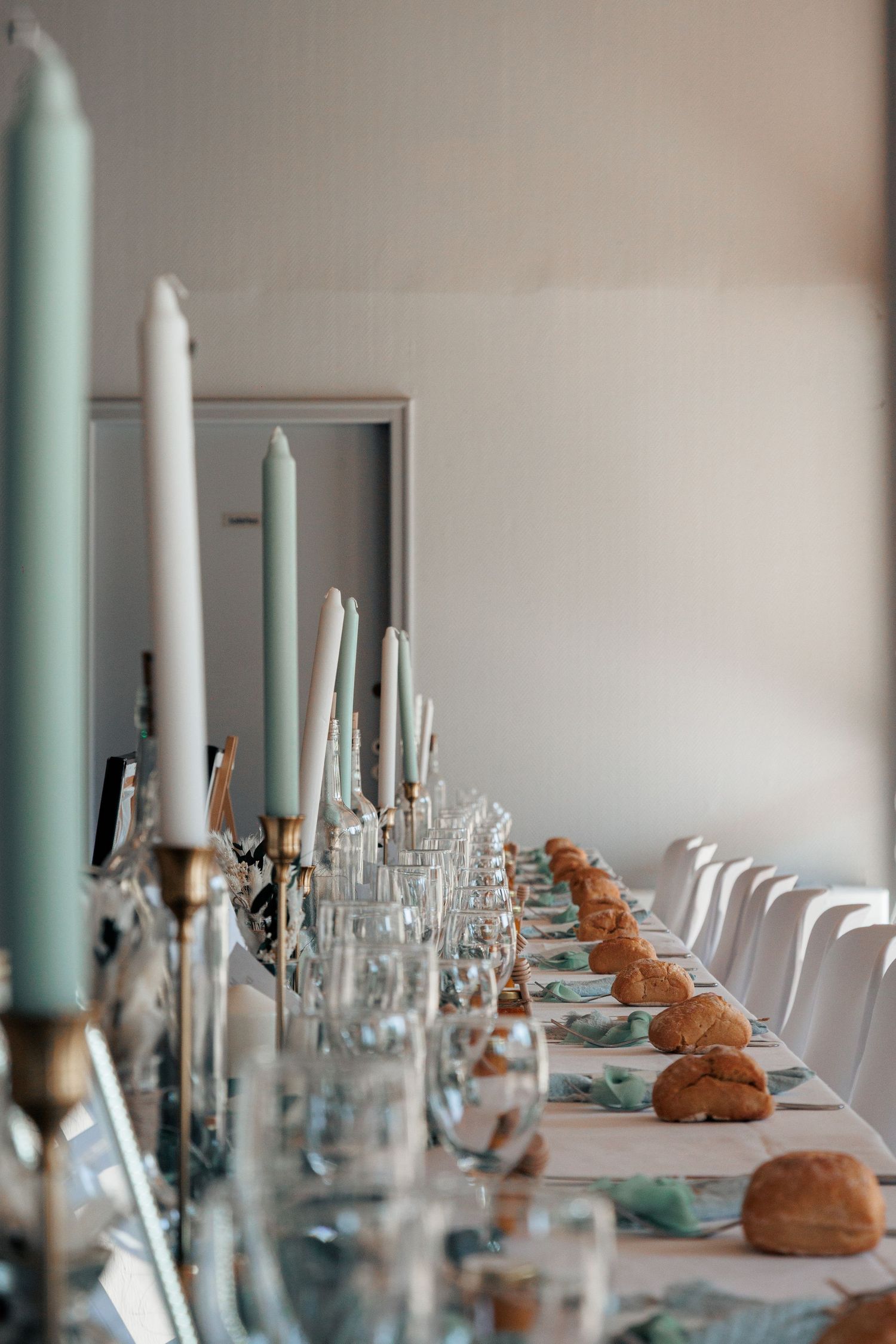 Long elegantly set wedding reception table with tall sage green and white candles, glassware, and bread rolls.