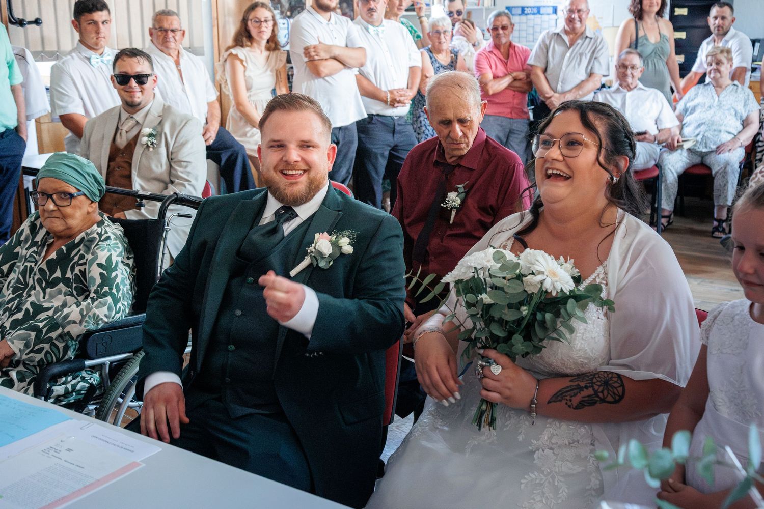 Smiling bride holding white bouquet and groom in teal suit sit together at wedding ceremony table with guests behind.