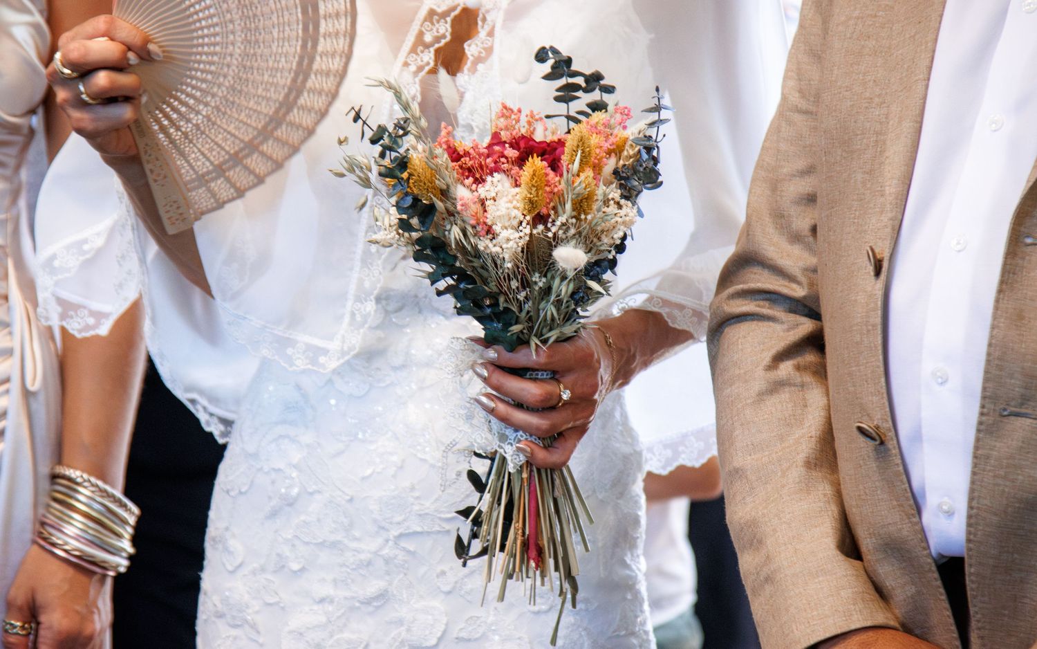 A wedding bouquet with dried flowers and fresh blooms in pastel orange and purple held between a white dress and tan suit.