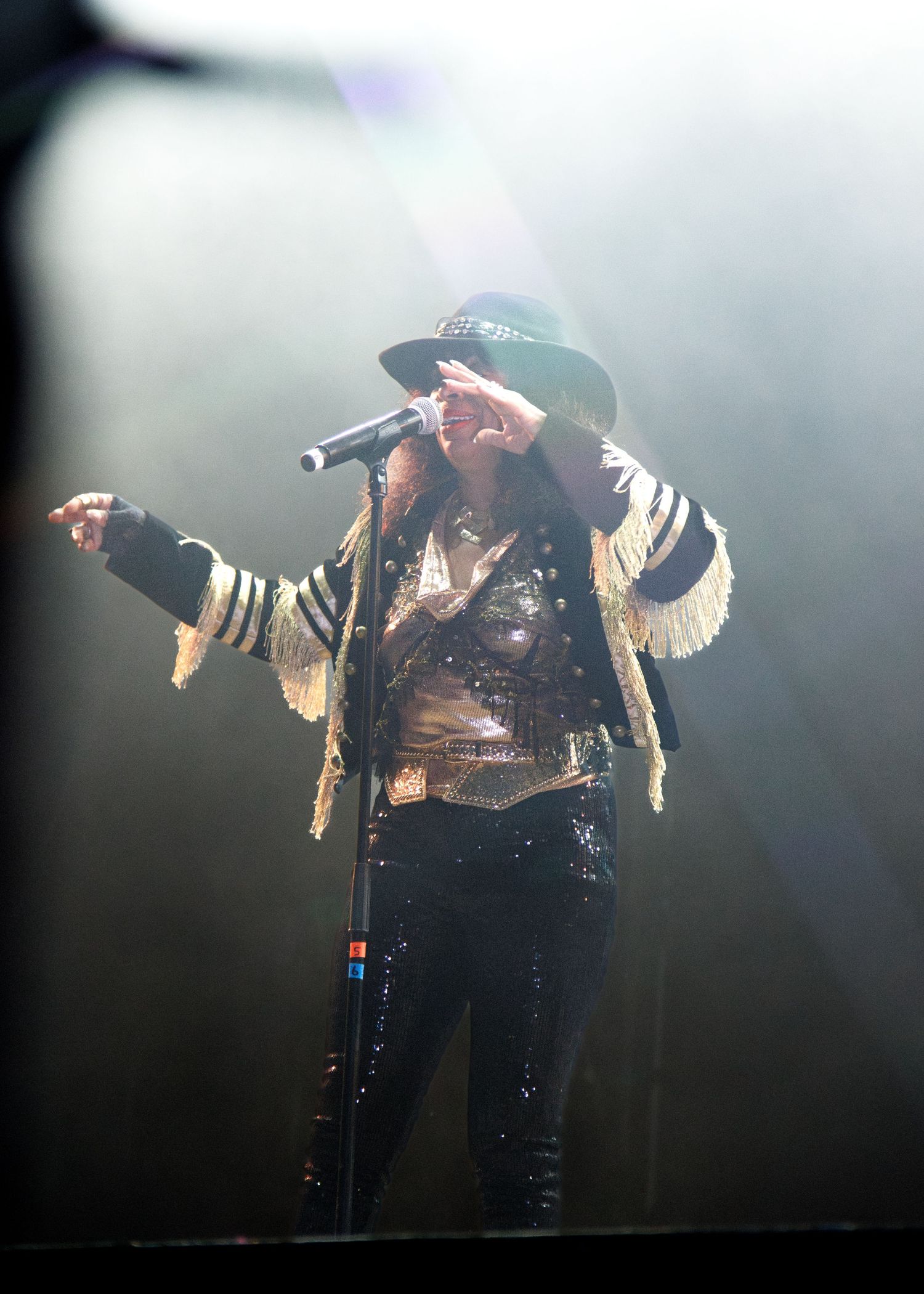 A performer in dark clothing with golden wings stands silhouetted on a smoky concert stage under dramatic lighting.