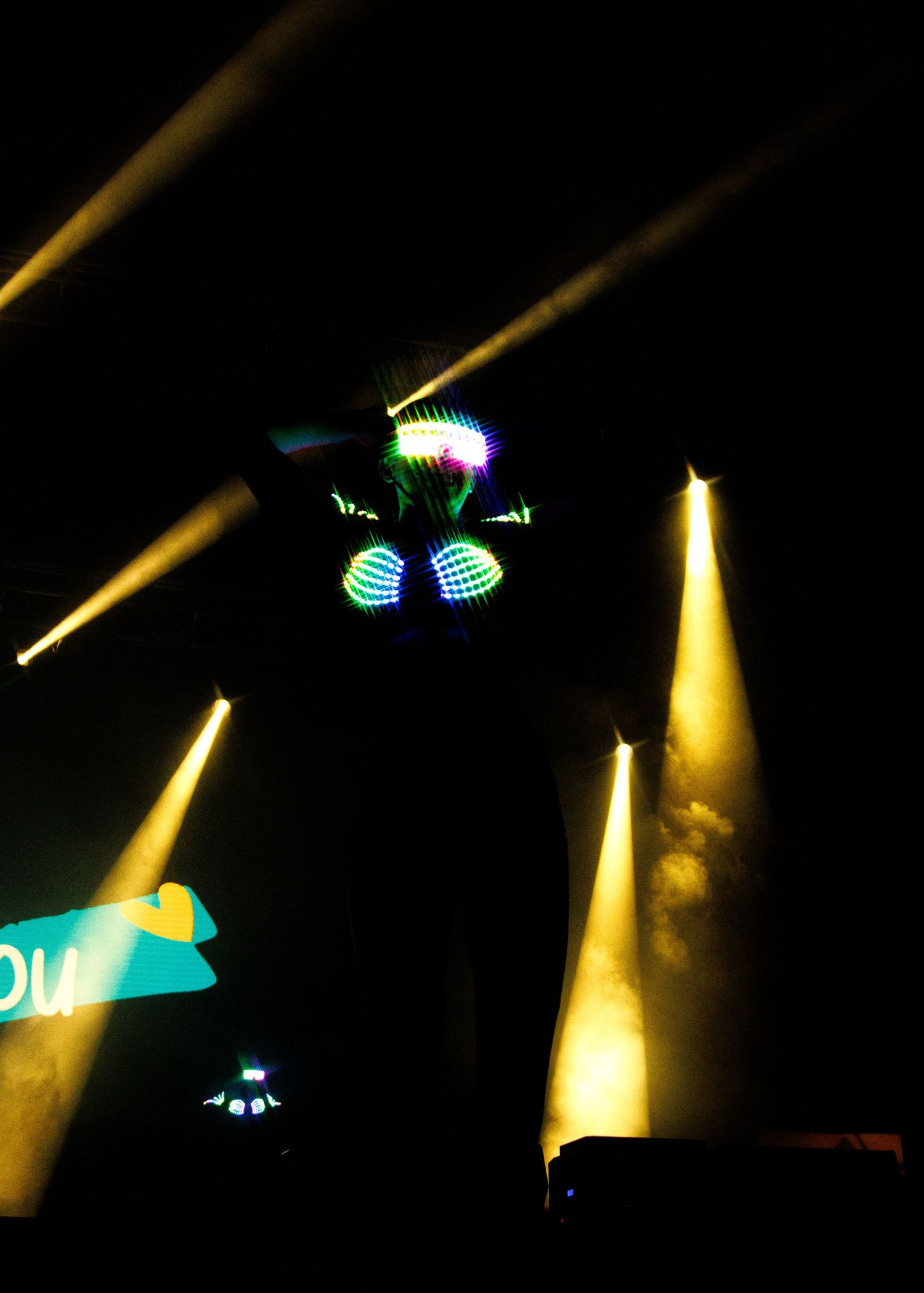 Dramatic yellow spotlights illuminate glowing neon blue elements in a dark nightclub atmosphere.